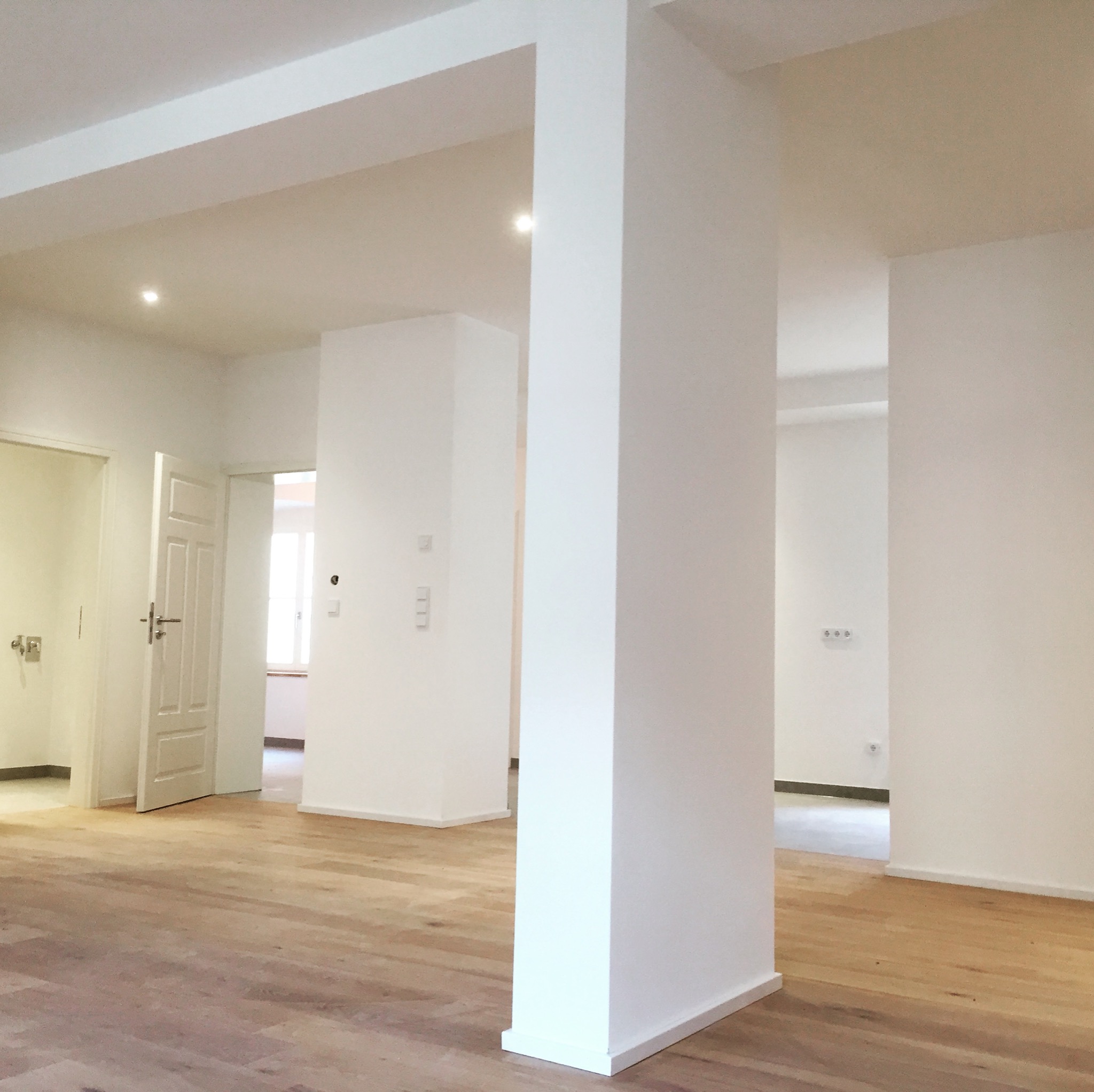Wnętrze nowo wykończonego, jasnego pomieszczenia z drewnianą podłogą, białymi ścianami, kolumnami nośnymi i wpuszczonym oświetleniem sufitowym. Widoczne otwarte drzwi prowadzące do kolejnych...