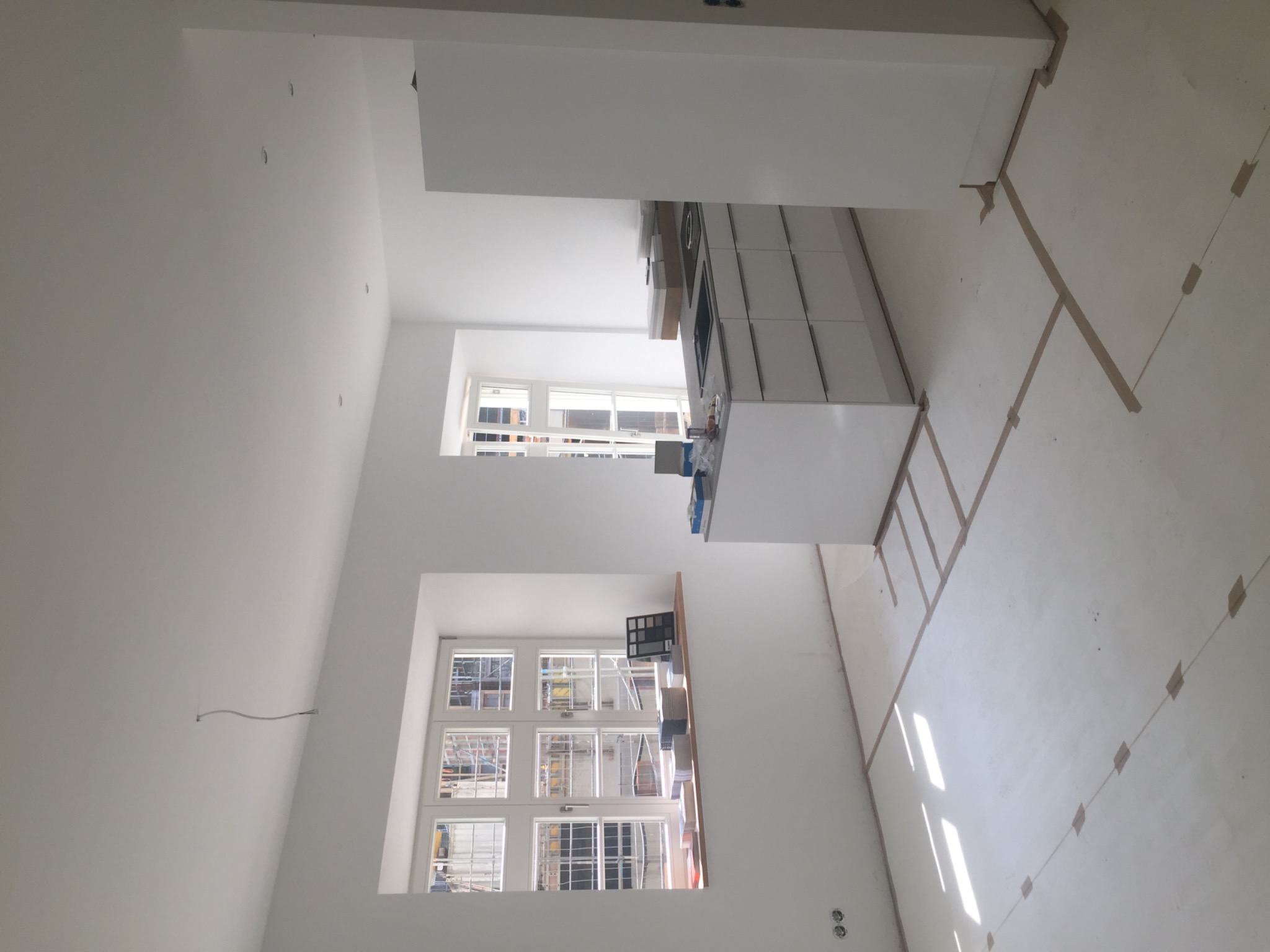 Wnętrze kuchni w trakcie remontu, widoczne przygotowania do instalacji oświetlenia, z przewodami wychodzącymi ze ściany, okna z widokiem na rusztowania, taśmy malarskie na podłodze wyznaczające...