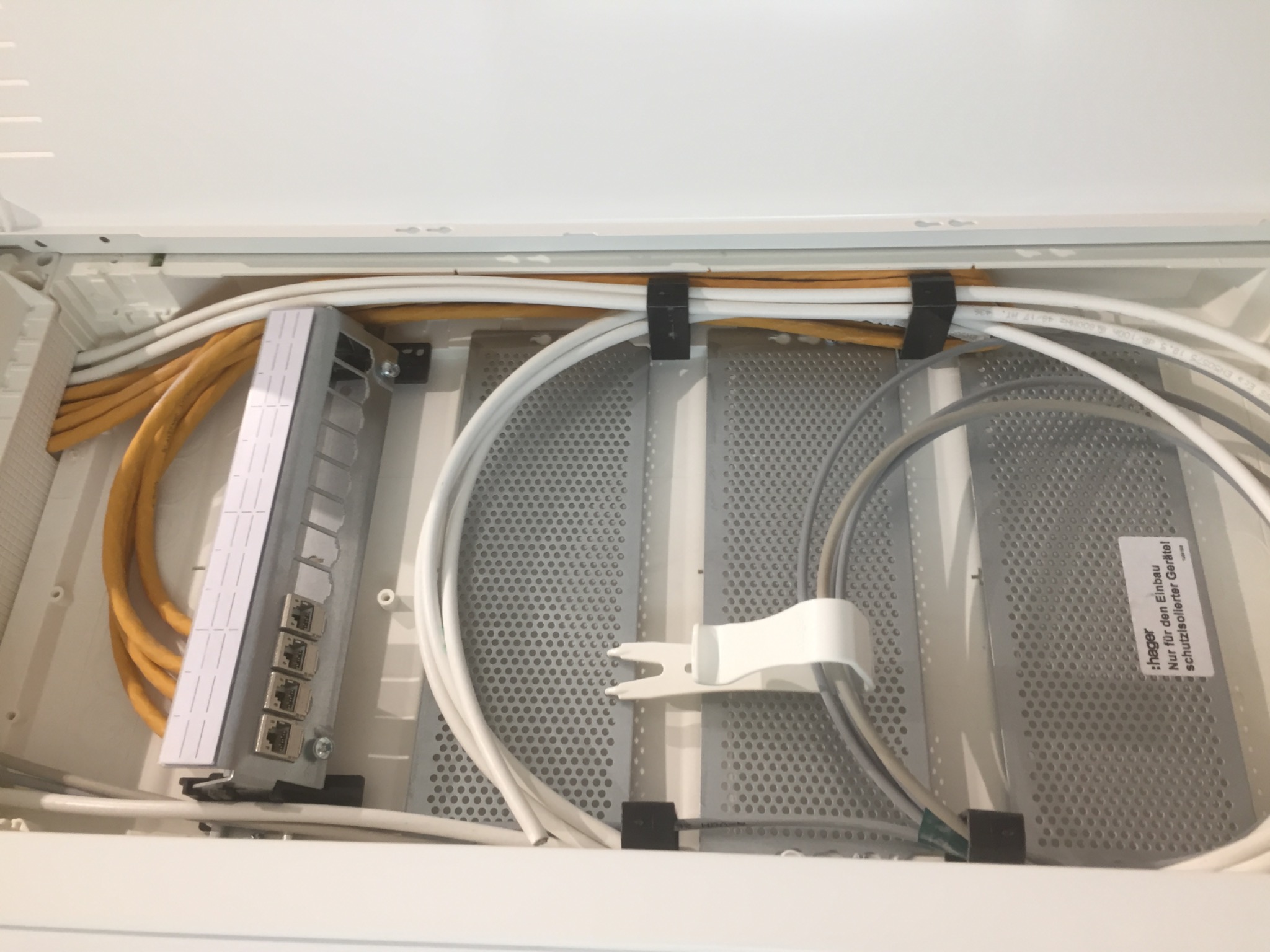 Wnętrze białej skrzynki rozdzielczej z panelami perforowanymi, zawierającej kable Ethernet w kolorach żółtym, białym i szarym, oraz metalowy panel krosowniczy z portami RJ45, zamocowane za pomocą...