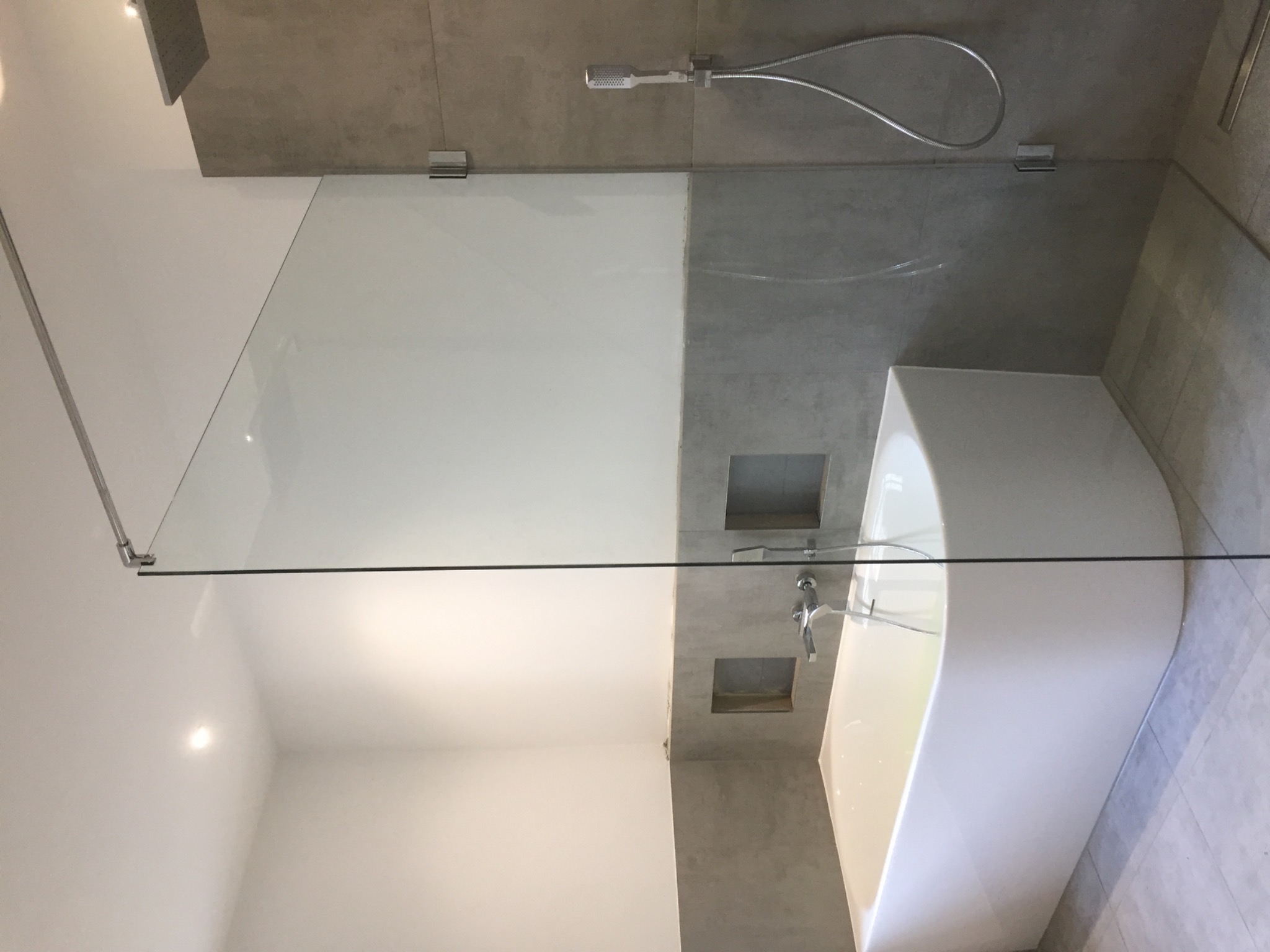 Nowoczesna łazienka z białą wanną i szklaną kabiną prysznicową, widoczne srebrne mocowania szkła i minimalistyczna armatura.