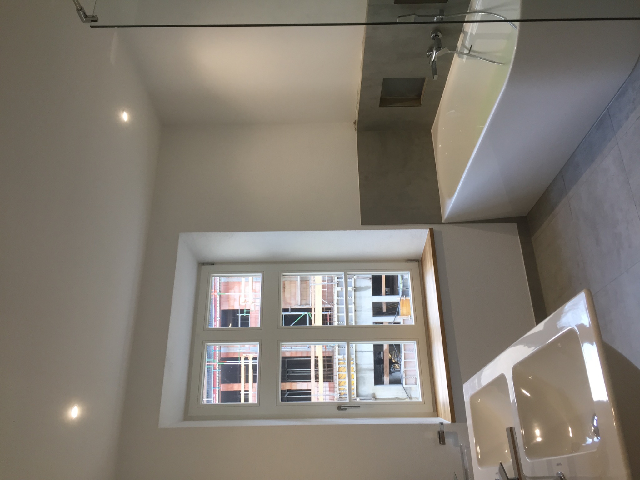 Nowoczesna łazienka w Gdańsku z białymi ścianami, widokiem na budowę przez okno, punktowe oświetlenie sufitowe, biała wanna i umywalka.