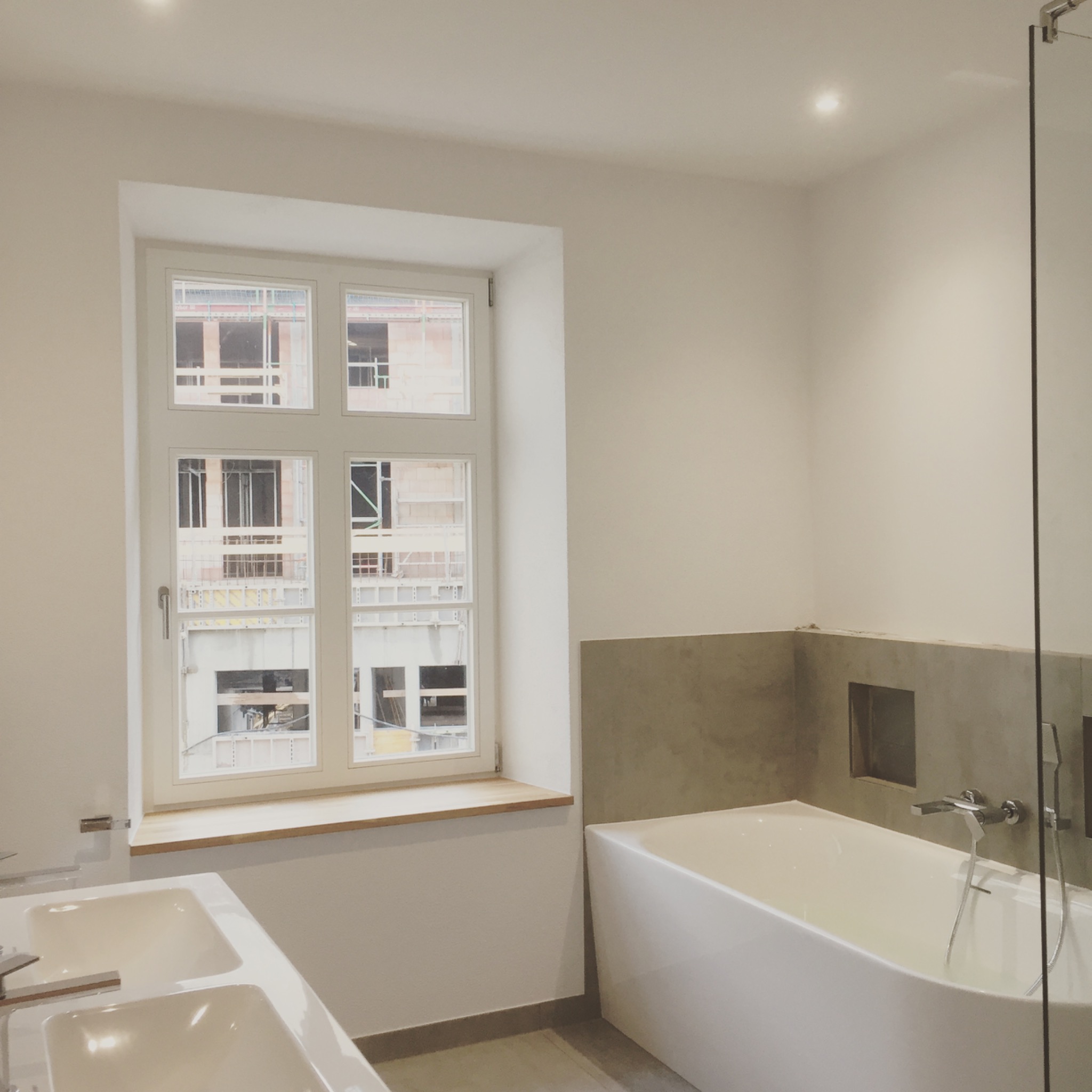 Nowoczesna łazienka z białymi ścianami, wanną z szarą obudową i podwójną umywalką. W oknie widoczny budynek w trakcie budowy.
