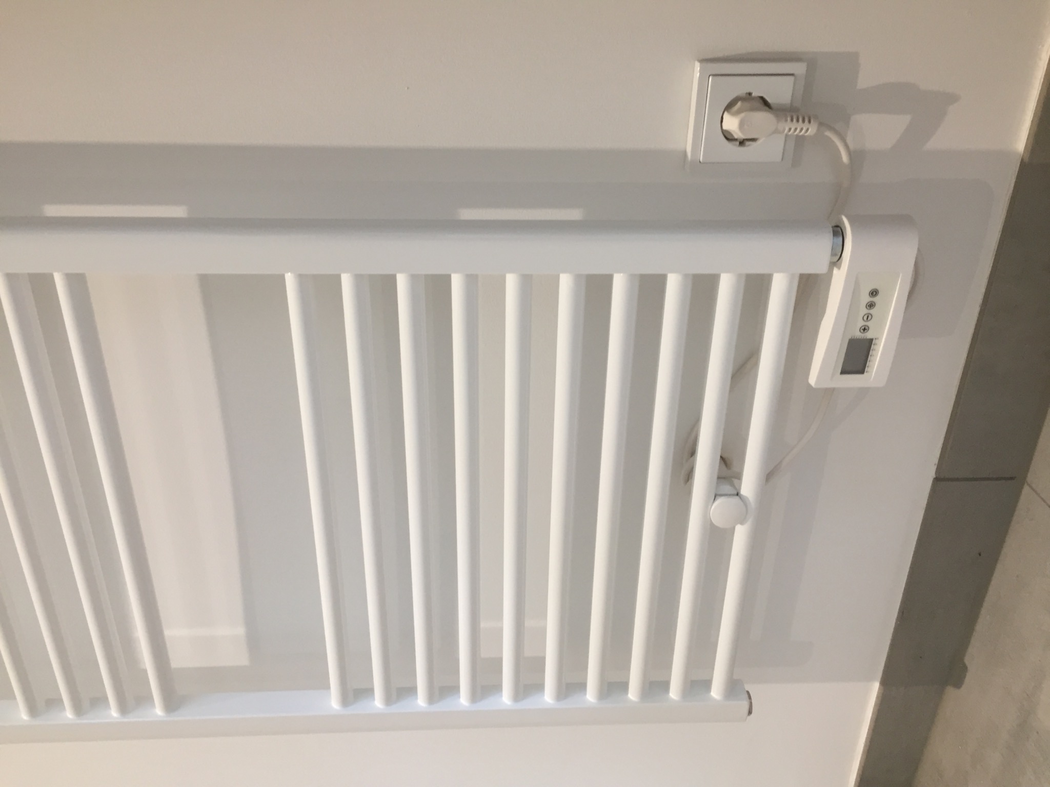 Biały, elektryczny grzejnik drabinkowy zamontowany na białej ścianie, podłączony do gniazdka elektrycznego z widocznym panelem sterowania.