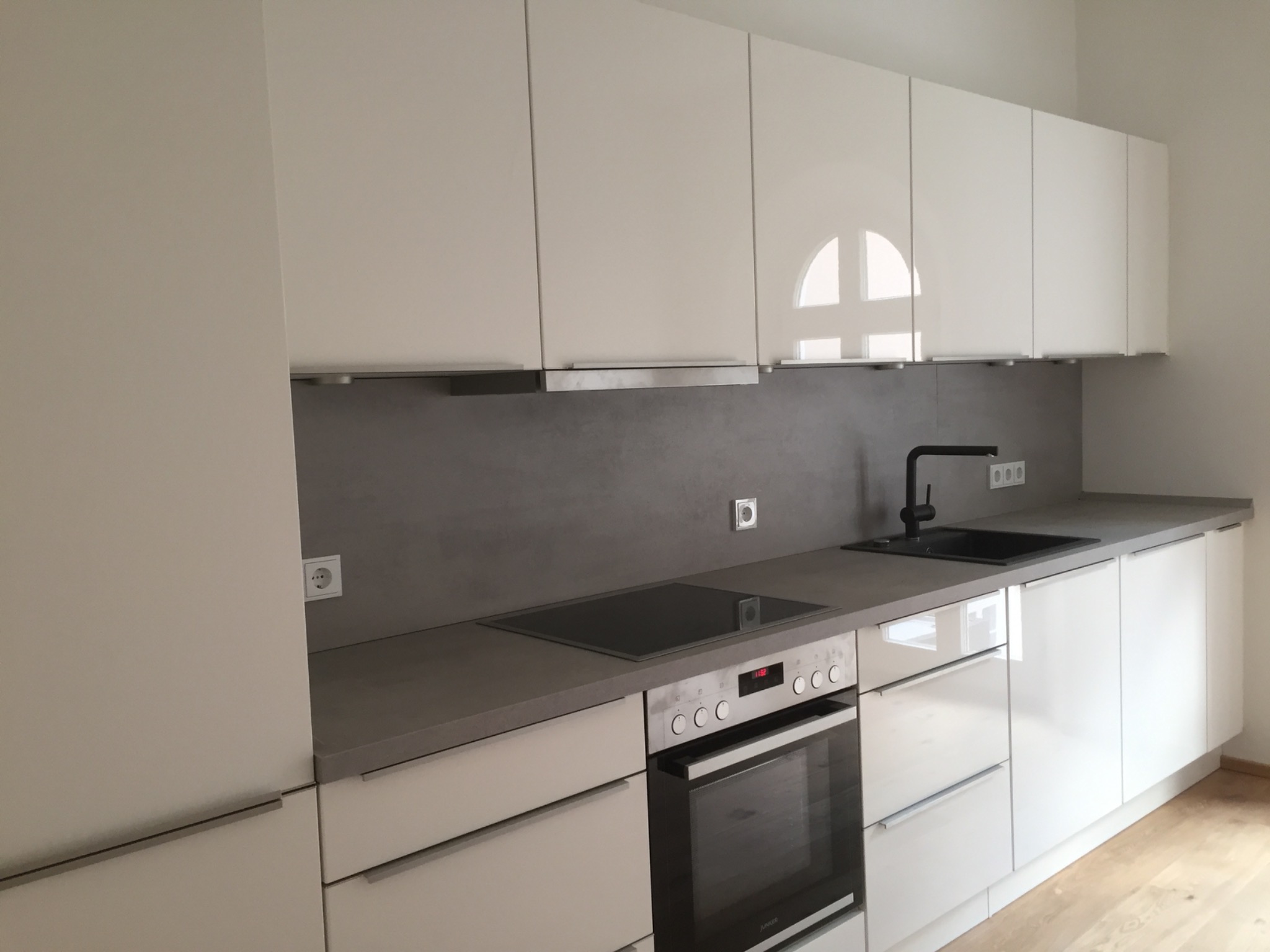 Nowoczesna, minimalistyczna kuchnia z białymi, lakierowanymi frontami szafek, szarym blatem i płytą indukcyjną. Widoczne gniazdko elektryczne nad blatem.