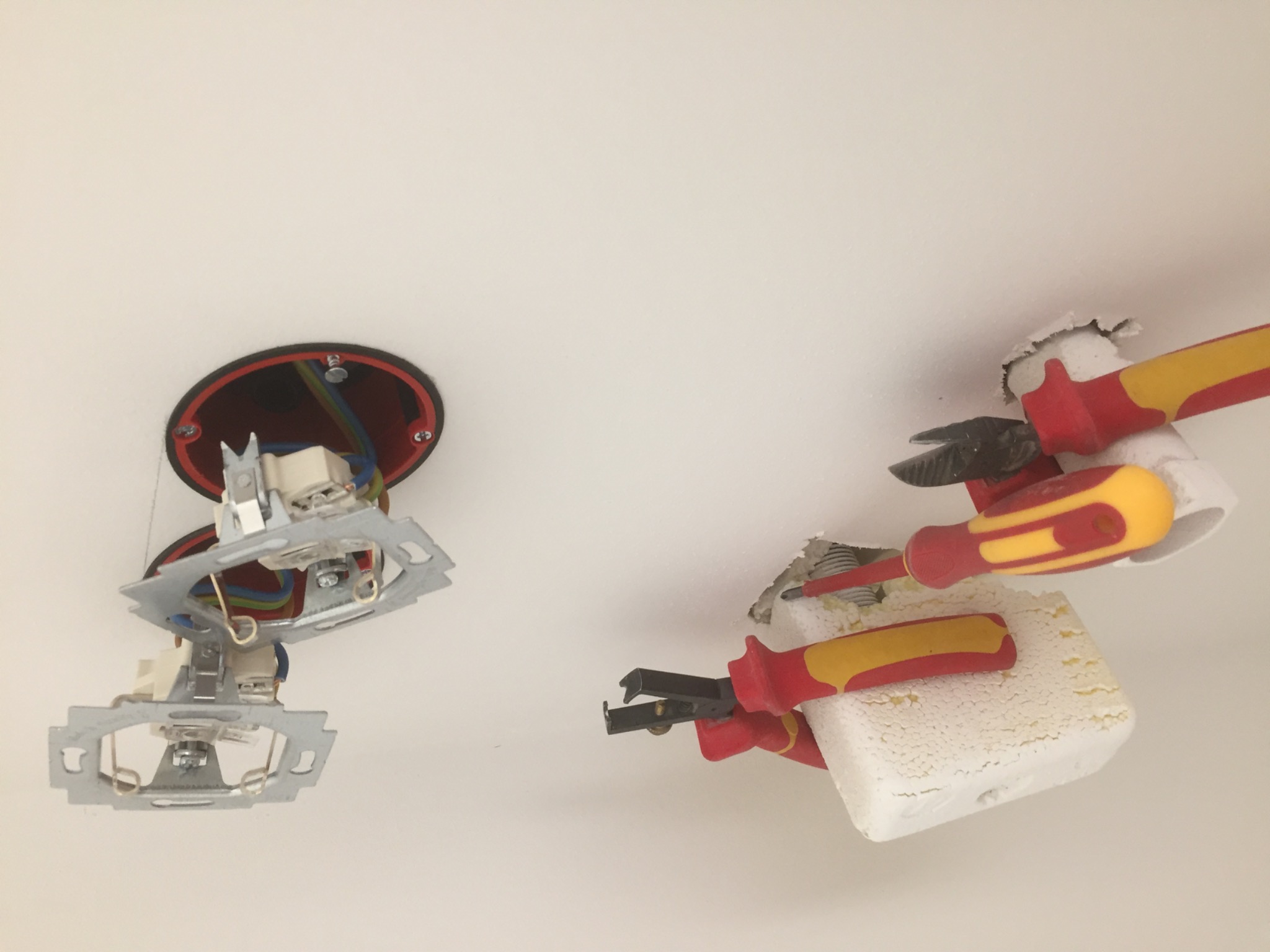 Widok z dołu na sufit podczas prac elektrycznych: widoczne kable, metalowe elementy mocujące, narzędzia (szczypce, śrubokręty) i kostka elektryczna z przewodami.