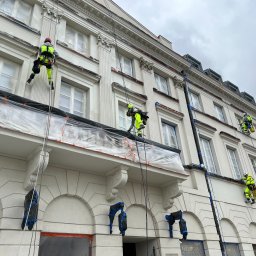CLEAN CITY SPÓŁKA CYWILNA - Pracownicy w jaskrawych kombinezonach na linach, zabezpieczają i malują elewację budynku z dekoracyjnymi elementami architektonicznymi. Okna zabezpieczone folią.