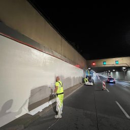 CLEAN CITY SPÓŁKA CYWILNA - Pracownik w żółtym kombinezonie natryskowo aplikuje farbę na ścianę tunelu drogowego. W tle widoczny podnośnik z innymi pracownikami i ruch uliczny.