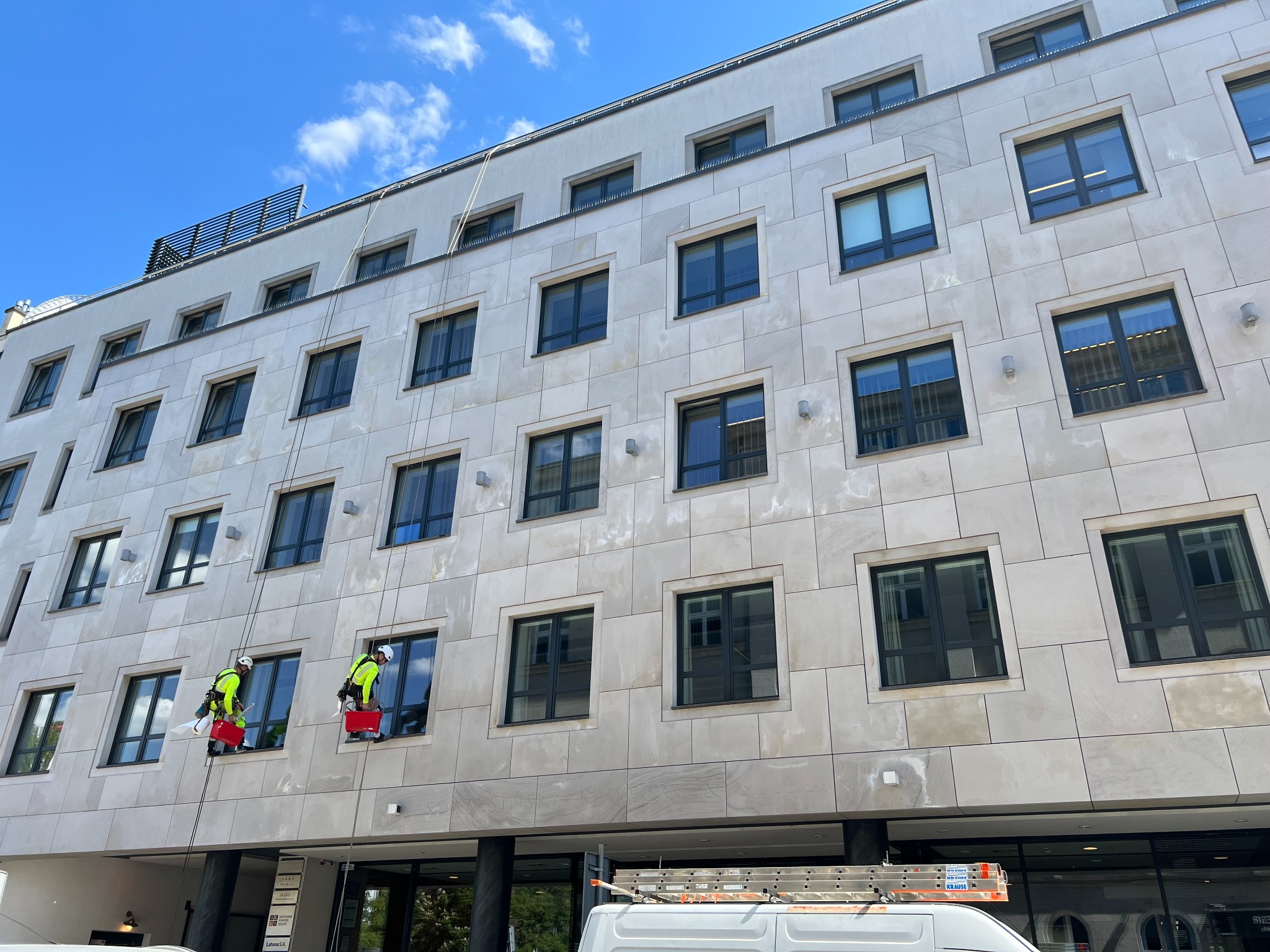 Dwóch pracowników w uprzężach czyści elewację budynku z jasnego kamienia. Widoczny fragment białego busa z drabiną. Błękitne niebo w tle.