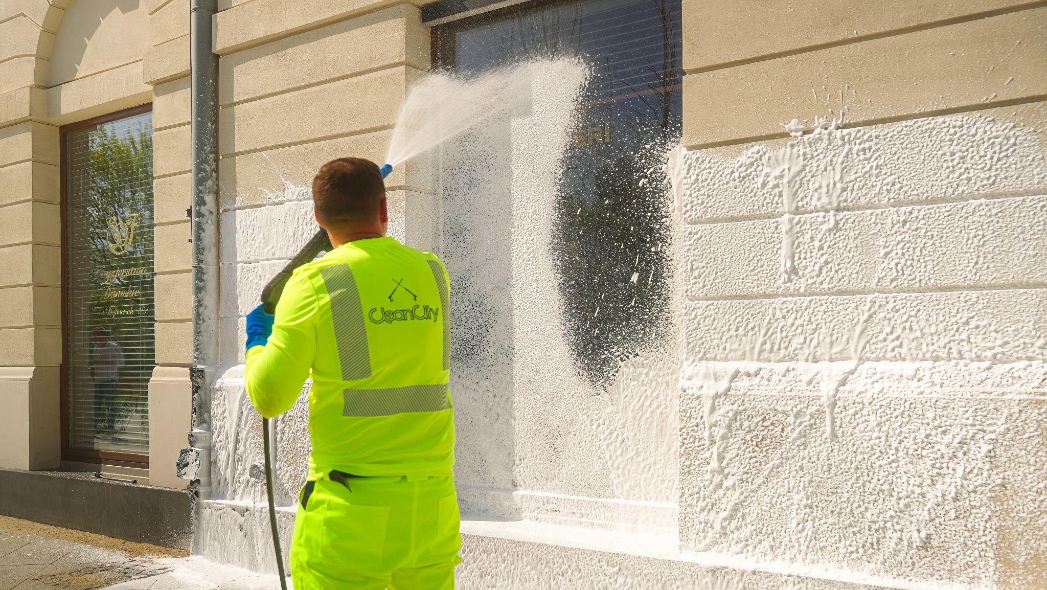 Pracownik w jaskrawożółtym kombinezonie czyści elewację budynku za pomocą myjki ciśnieniowej, pokrywając ją białą pianą. Widoczny fragment okna z żaluzjami.