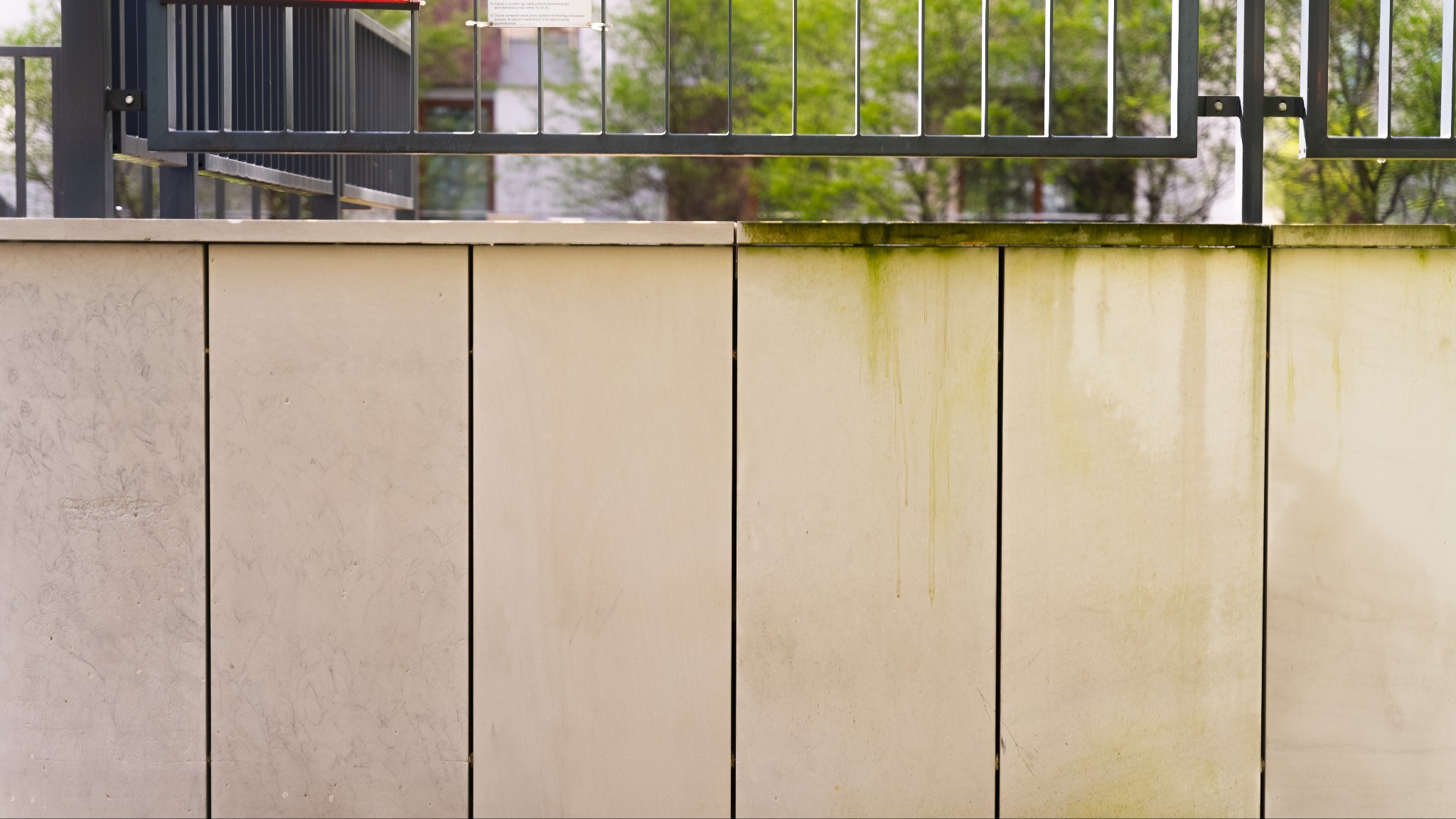 Fragment kamiennej elewacji z widocznymi śladami zabrudzeń i porostów, kontrastujący z czystymi panelami po lewej stronie. Metalowe ogrodzenie w tle.