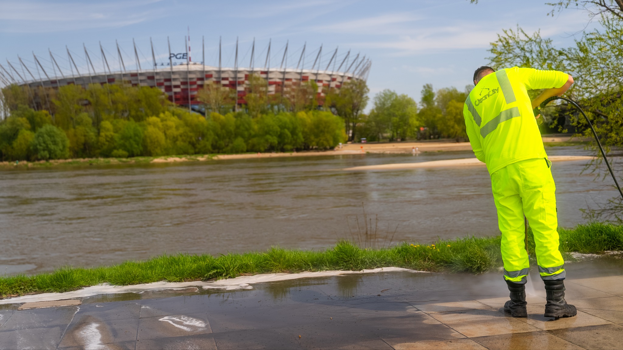 Pracownik w jaskrawożółtym stroju czyści chodnik myjką ciśnieniową. W tle rzeka, zieleń i Stadion Narodowy w Warszawie, w słoneczny dzień.