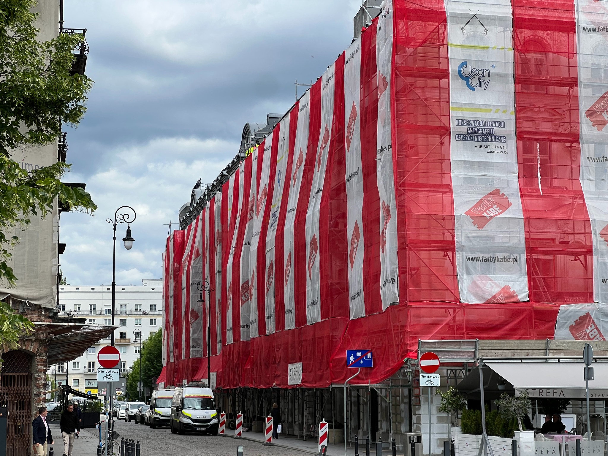 Budynek w rusztowaniach, osłonięty siatką z reklamami firm remontowych. Widok z ulicy, fragment chodnika i ulicy z zaparkowanymi autami. Pochmurne niebo.