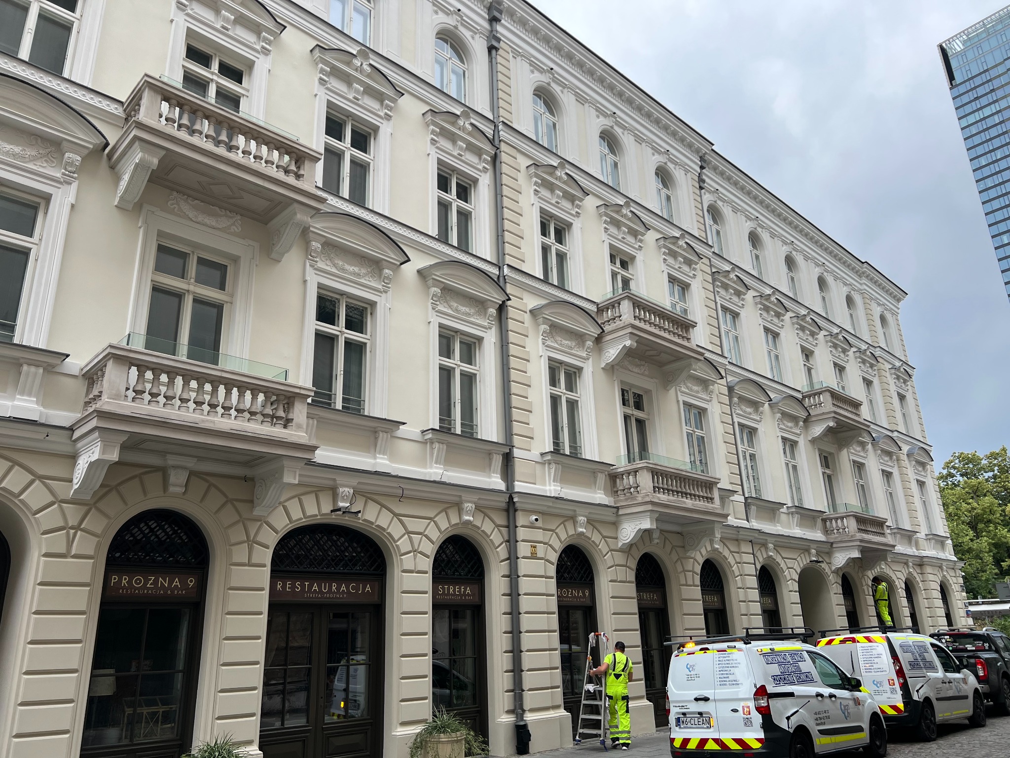 Pracownik w jaskrawym kombinezonie na drabinie, myjący elewację kamienicy z restauracjami. W tle zaparkowane busy firmy sprzątającej.