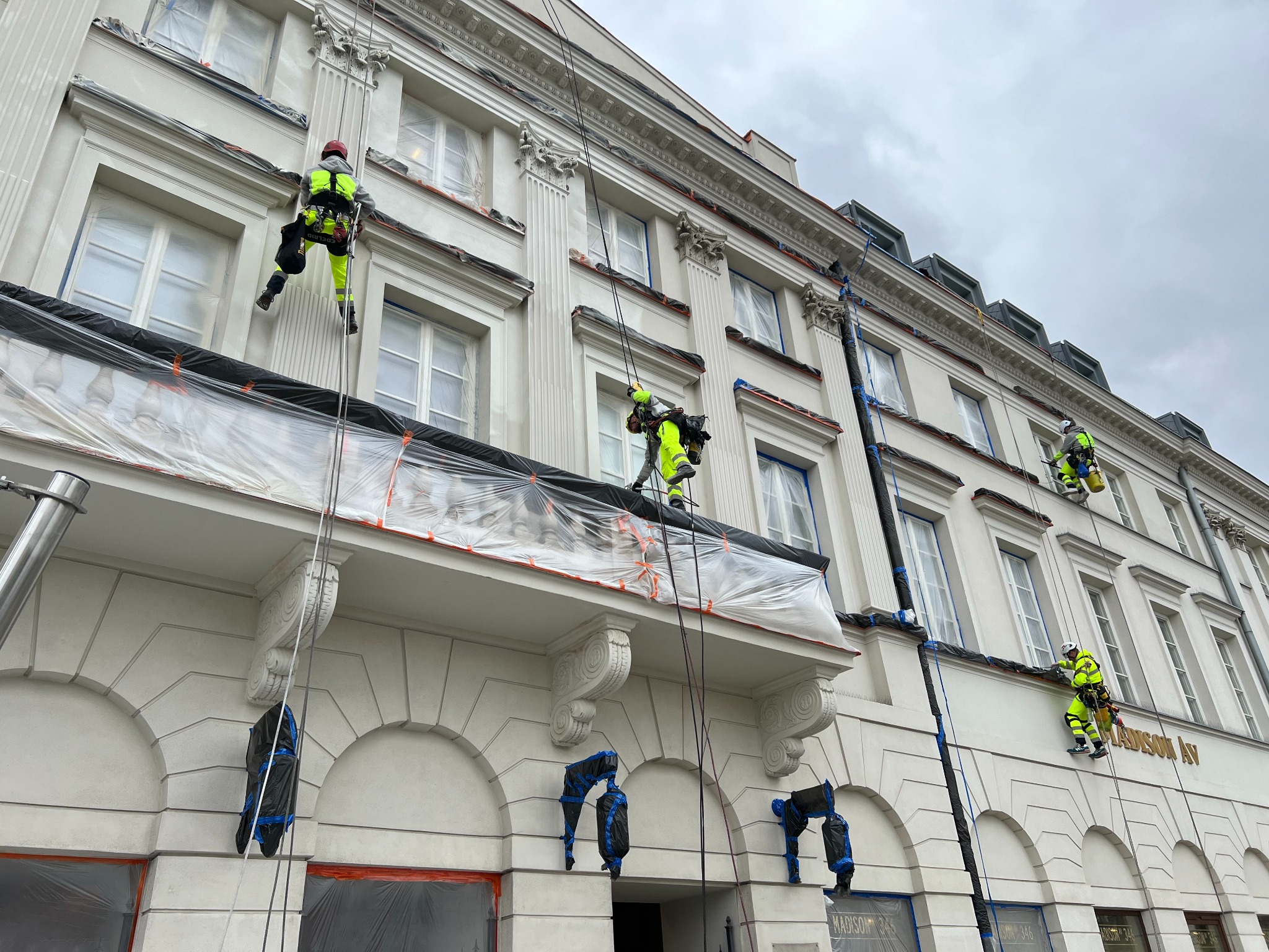Pracownicy w jaskrawych kombinezonach na linach, zabezpieczają i malują elewację budynku z dekoracyjnymi elementami architektonicznymi. Okna zabezpieczone folią.