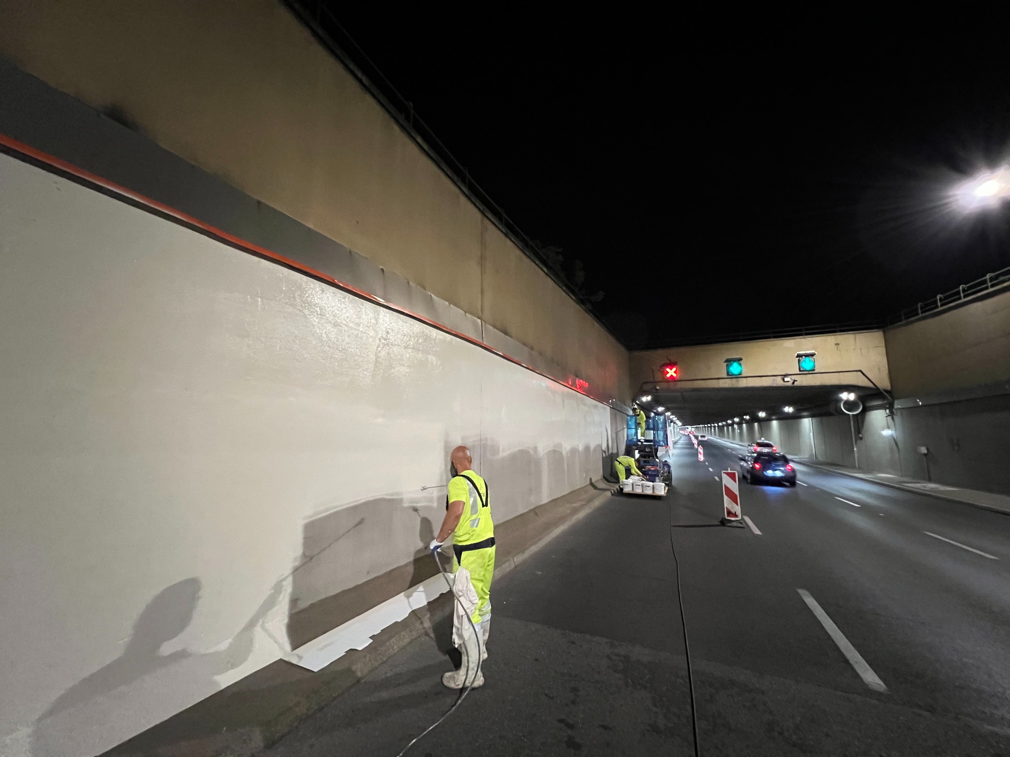 Pracownik w żółtym kombinezonie natryskowo aplikuje farbę na ścianę tunelu drogowego. W tle widoczny podnośnik z innymi pracownikami i ruch uliczny.