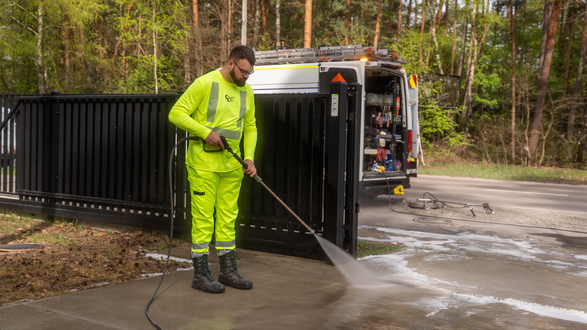 Pracownik w odblaskowym stroju czyści chodnik myjką ciśnieniową. W tle brama, ulica i otwarty samochód dostawczy z narzędziami. Widoczne efekty czyszczenia.
