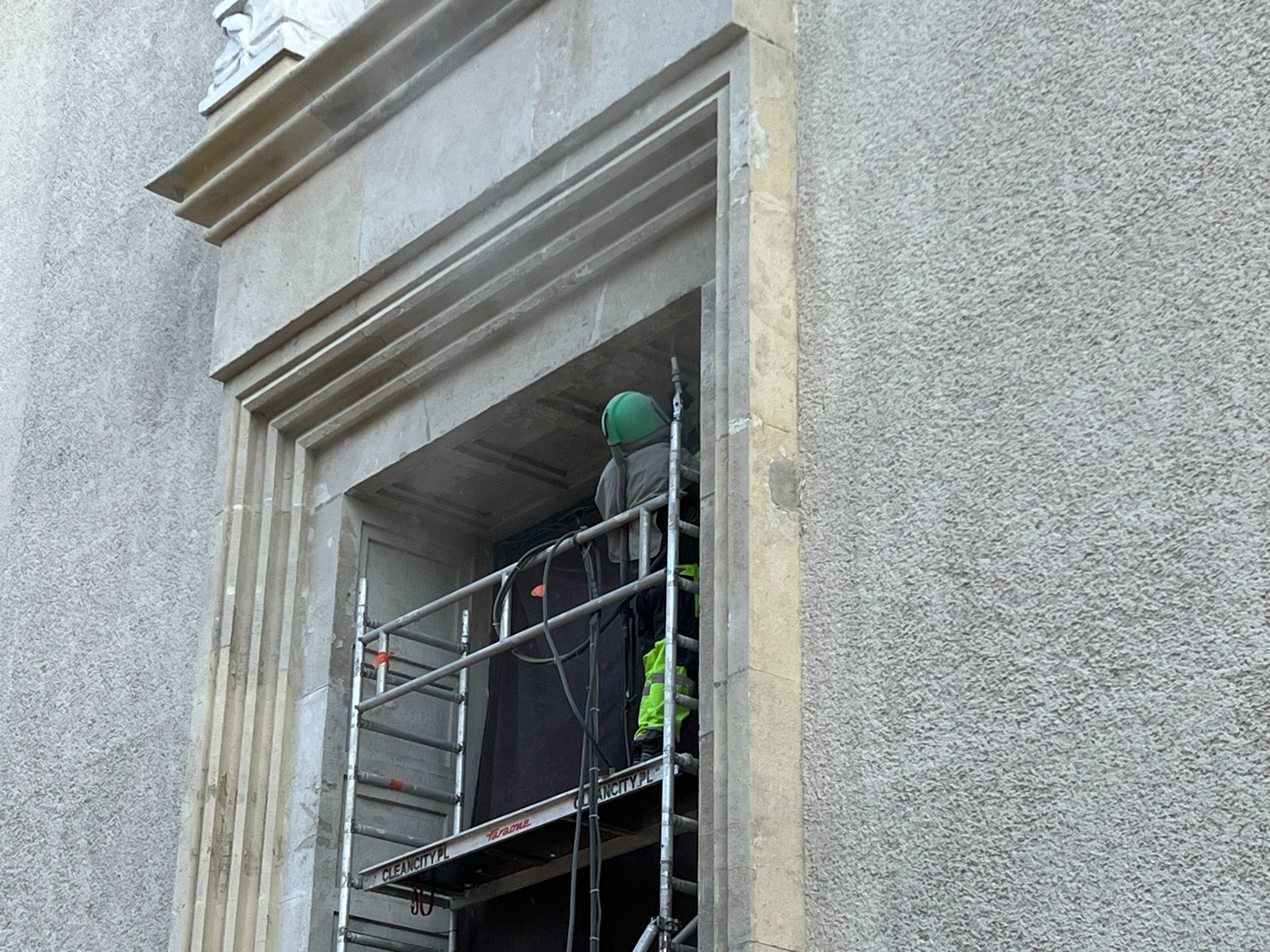 Pracownik w kasku na rusztowaniu czyści kamienną elewację budynku. Widoczne detale architektoniczne i struktura elewacji. Ujęcie z bliska, skupione na detalu.
