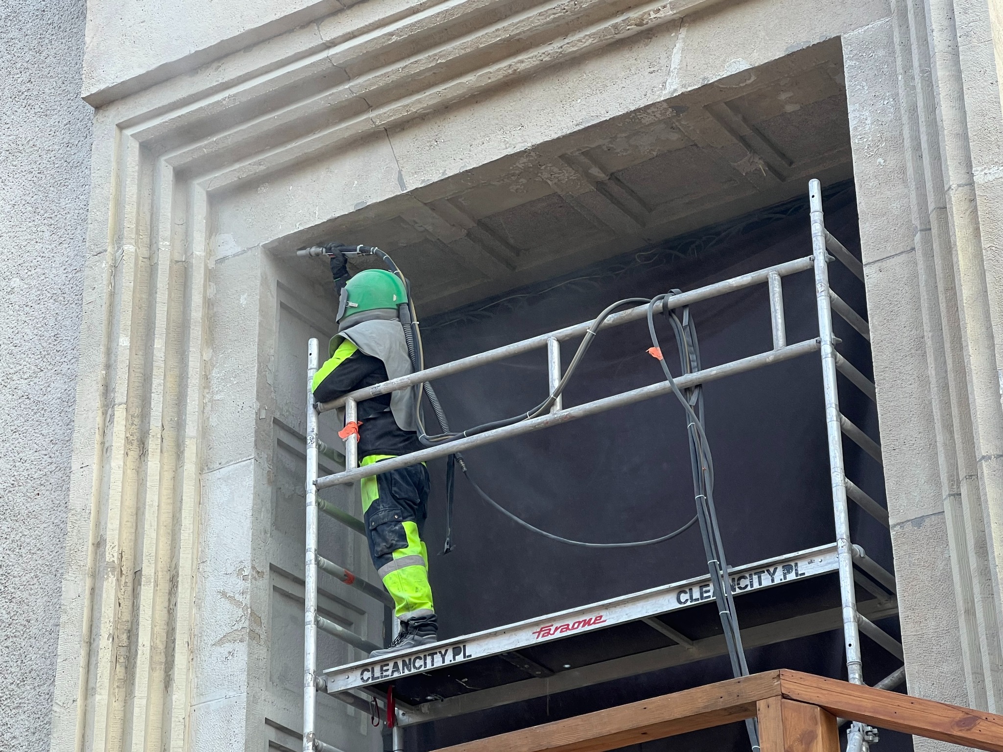 Pracownik w kombinezonie ochronnym na rusztowaniu, piaskuje kamienną elewację budynku. Widoczne detale architektoniczne i narzędzie pracy.