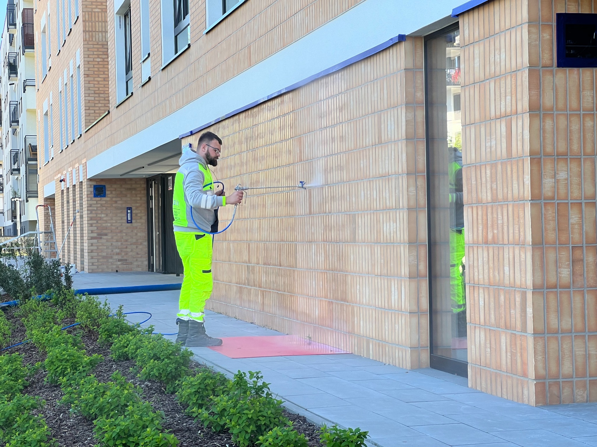 Pracownik w odblaskowym stroju impregnuje ceglaną elewację budynku za pomocą pistoletu natryskowego. Widoczne zabezpieczenia roślin i chodnika.