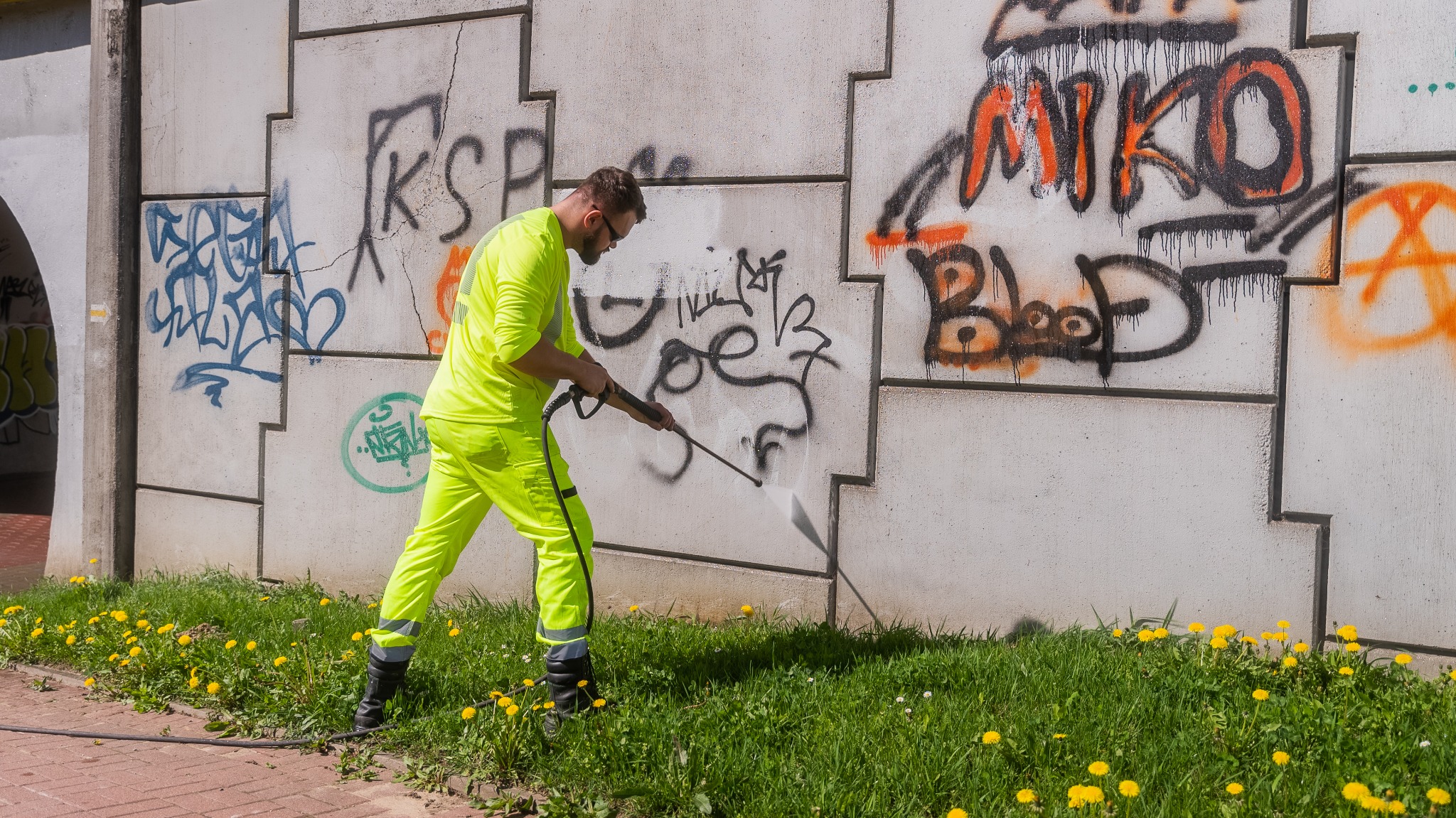 Pracownik w jaskrawożółtym kombinezonie usuwa graffiti z betonowego muru za pomocą myjki ciśnieniowej. W tle widać tunel i zieleń z żółtymi kwiatami.