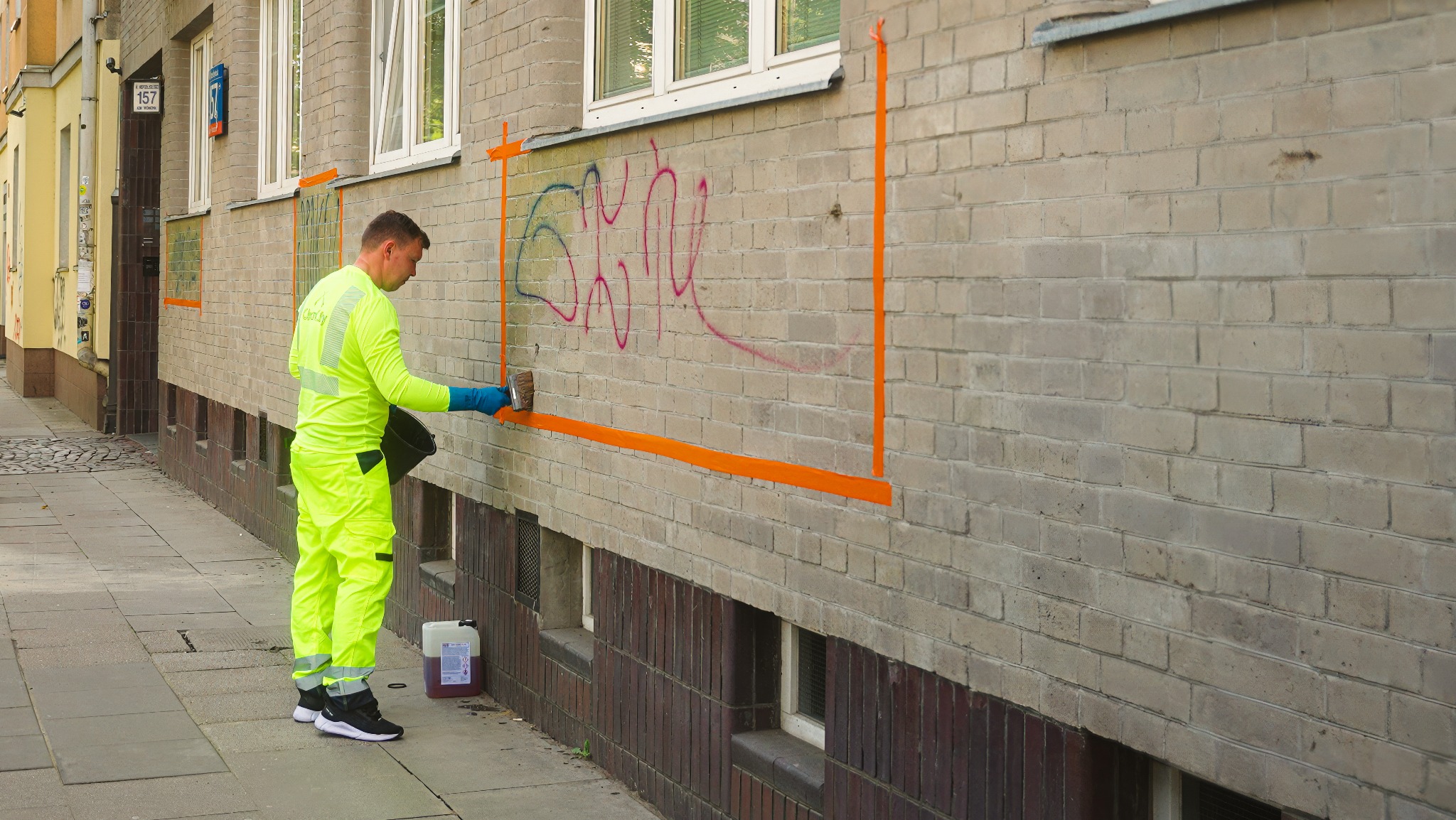 Pracownik w jaskrawożółtym stroju usuwa graffiti z ceglanej ściany budynku, używając pędzla i środka czyszczącego. Obszar pracy oznaczony pomarańczową taśmą.