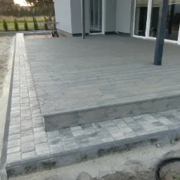 Szary taras z desek, otoczony kostką brukową, w trakcie budowy lub wykończenia, widoczne elementy konstrukcyjne i niezabudowany teren wokół.