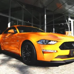 Pomarańczowy Ford Mustang zaparkowany przed budynkiem z przeszkloną fasadą, widoczny również fragment niebieskiego samochodu w tle.