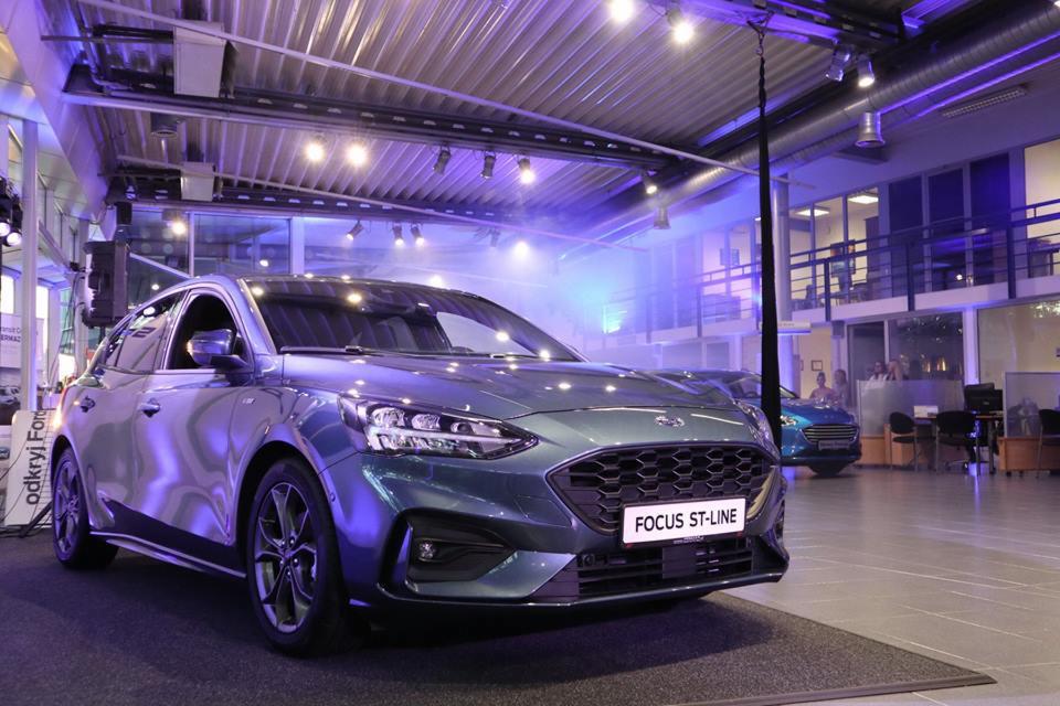 Zielony Ford Focus ST-Line na ekspozycji w salonie samochodowym, oświetlony reflektorami, z widocznym napisem 'odkryj Forda' na plakacie w tle.