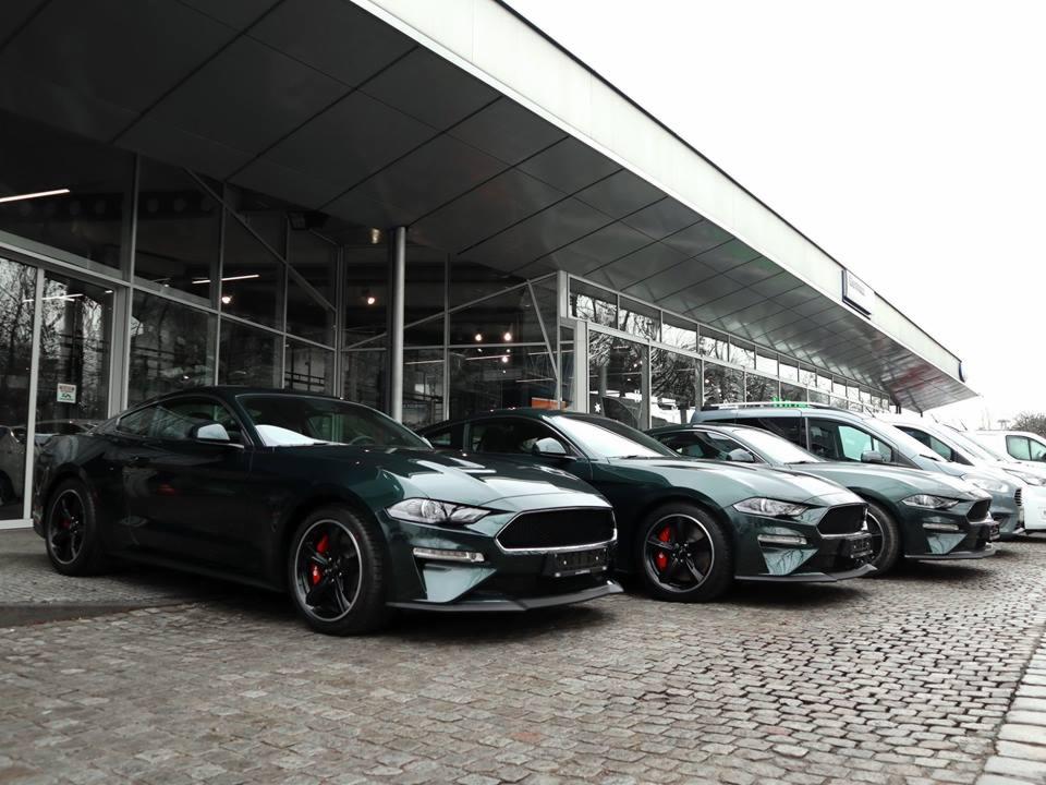 Szereg ciemnozielonych Fordów Mustangów zaparkowanych przed nowoczesnym salonem samochodowym z przeszkloną fasadą i szarym brukowanym chodnikiem.