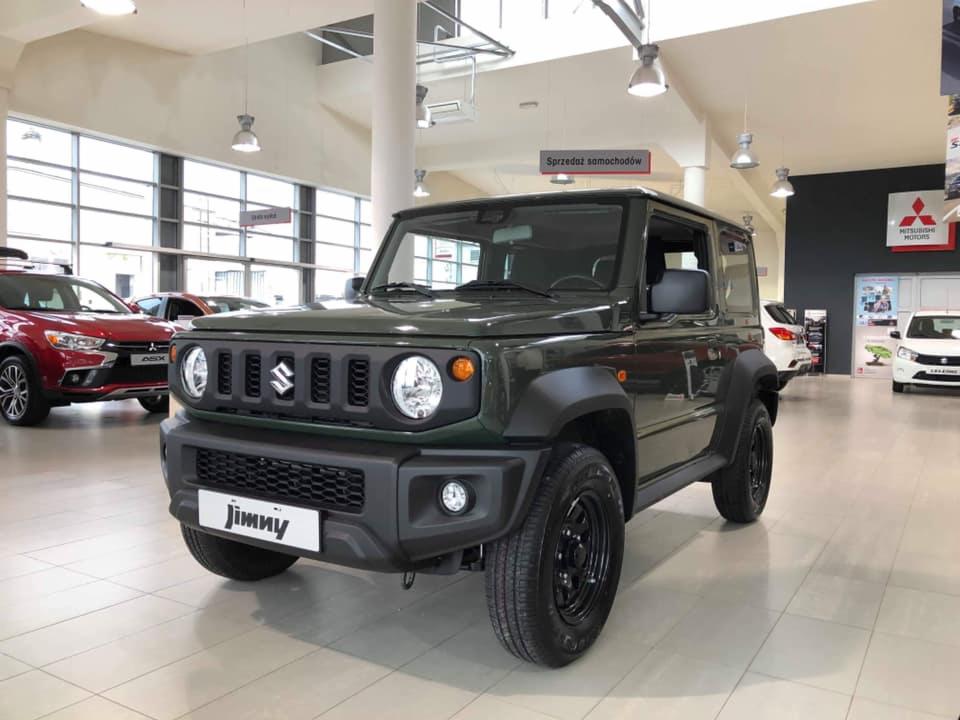 Nowy, zielony Suzuki Jimny prezentowany w salonie samochodowym, z widocznymi innymi modelami w tle i tablicą informacyjną 'Sprzedaż samochodów' pod sufitem.