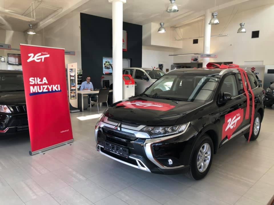 Czarny samochód osobowy marki Mitsubishi Outlander, udekorowany czerwoną wstążką i logotypem Radia ZET, prezentowany w salonie samochodowym na jasnej, kafelkowej podłodze. W tle widoczny baner...