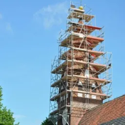 Wieża kościoła w trakcie renowacji, otoczona metalowymi rusztowaniami z drewnianymi pomostami, widoczne czerwone dachówki i fragment zielonego drzewa na tle błękitnego nieba.