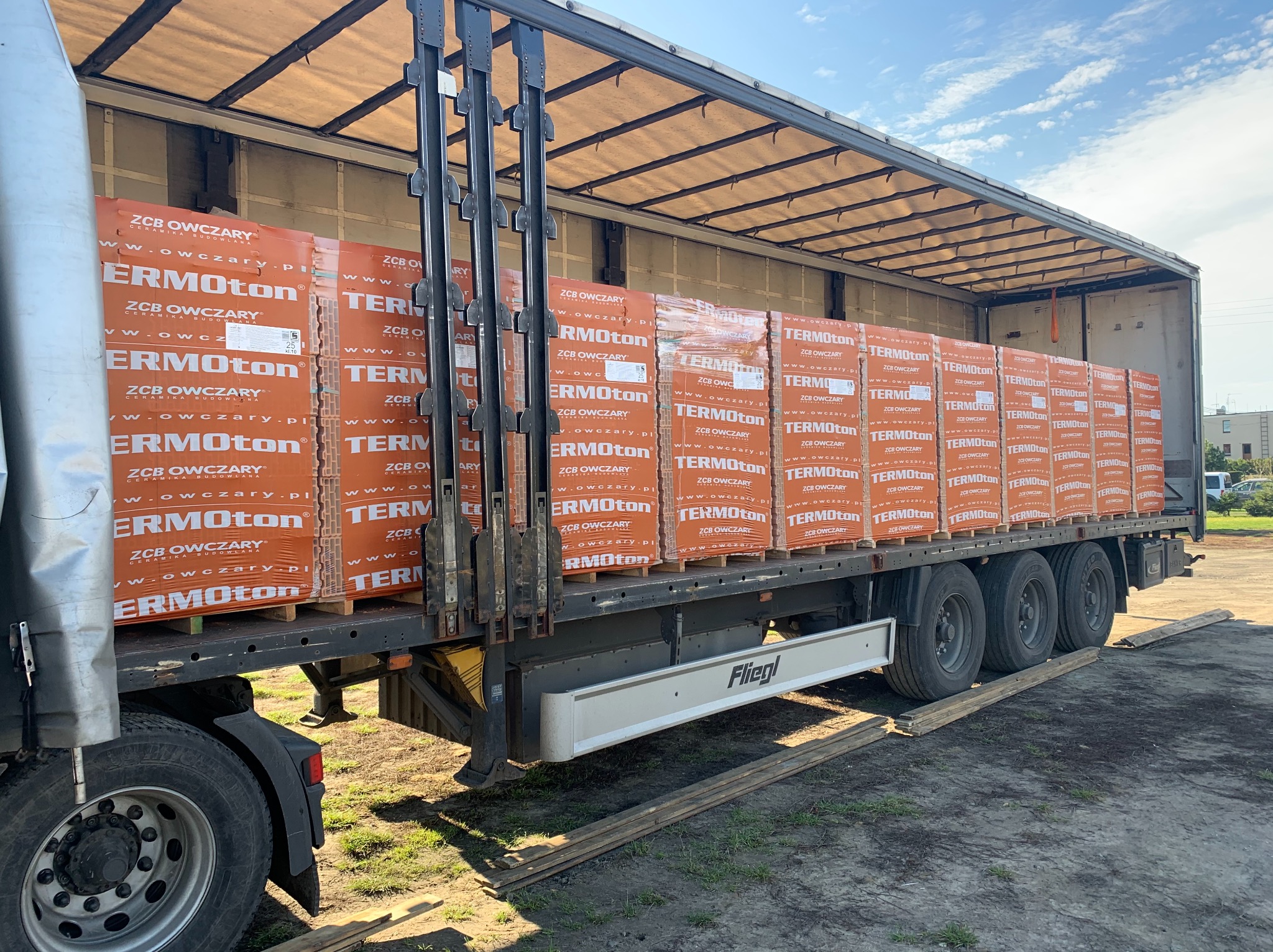 Ciężarówka z naczepą załadowana paletami z cegłami ceramicznymi Termoton, widok z boku na koła i dolną część naczepy, pod spodem leżą drewniane deski.