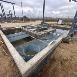 Budownictwo - inż. Jakub Kasica - Wykop fundamentowy z betonowymi ścianami, metalowymi kratami i konstrukcją stalową. W tle widoczny płot z drutu kolczastego i pochmurne niebo.