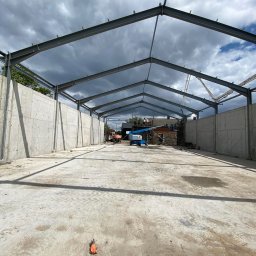 Budownictwo - inż. Jakub Kasica - Konstrukcja hali z betonu i stali w Kutnie, widok wnętrza. Na pierwszym planie betonowa posadzka, w tle maszyny budowlane i pochmurne niebo.