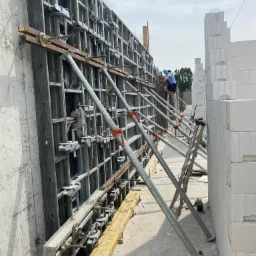 Szare szalunki wsparte metalowymi stemplami przy betonowej ścianie i murze z bloczków, dwóch robotników w oddali.