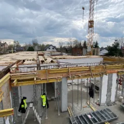 Wysoki kąt widzenia na plac budowy z widocznymi elementami konstrukcyjnymi, szalunkami, zbrojeniami i pustakami ceramicznymi, z dźwigiem w tle i pochmurnym niebem.
