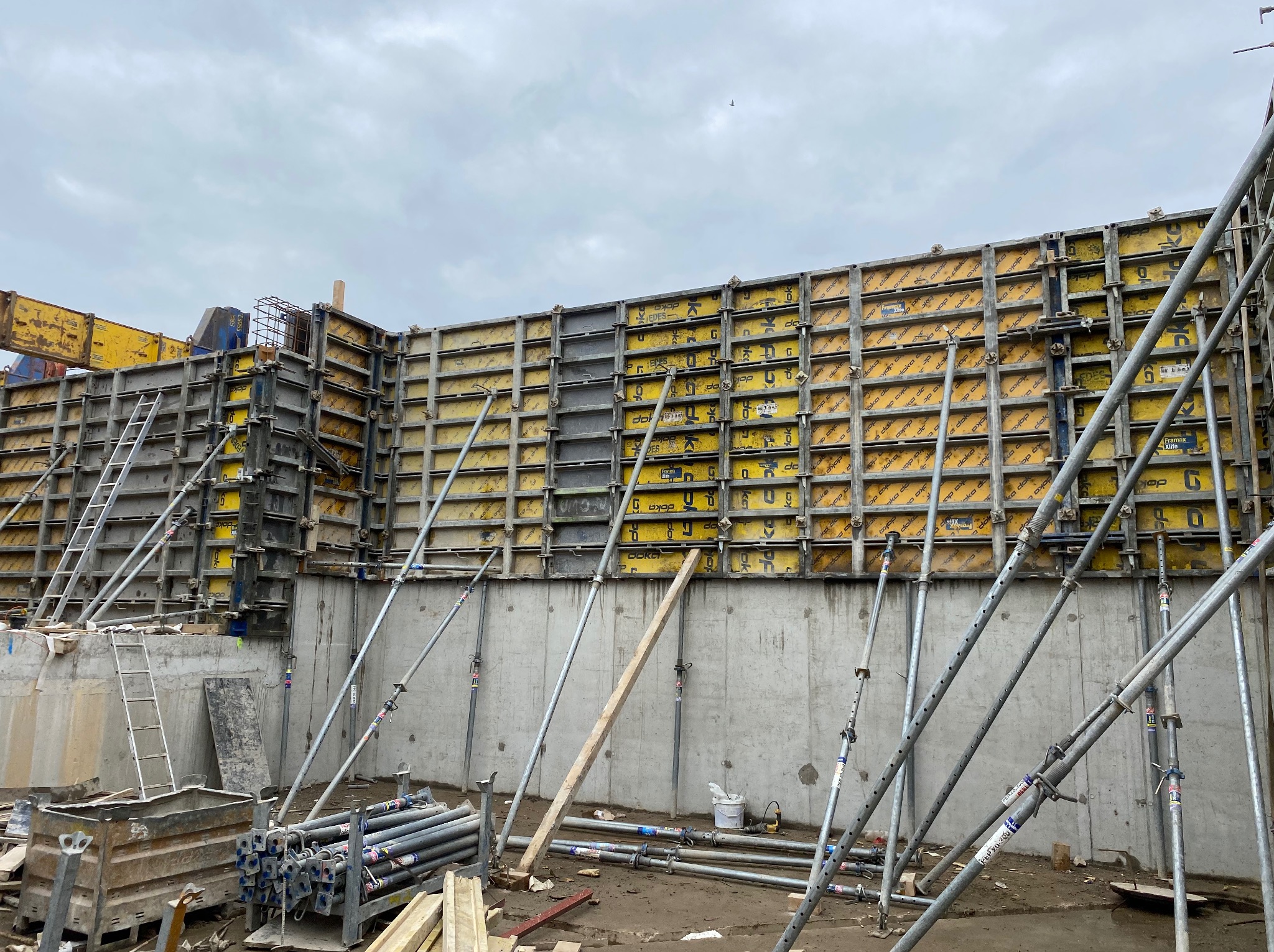 Szare szalunki Doka z żółtymi panelami, wsparte metalowymi stemplami, tworzą formę fundamentu pod budynek. Widoczne narzędzia i materiały budowlane.