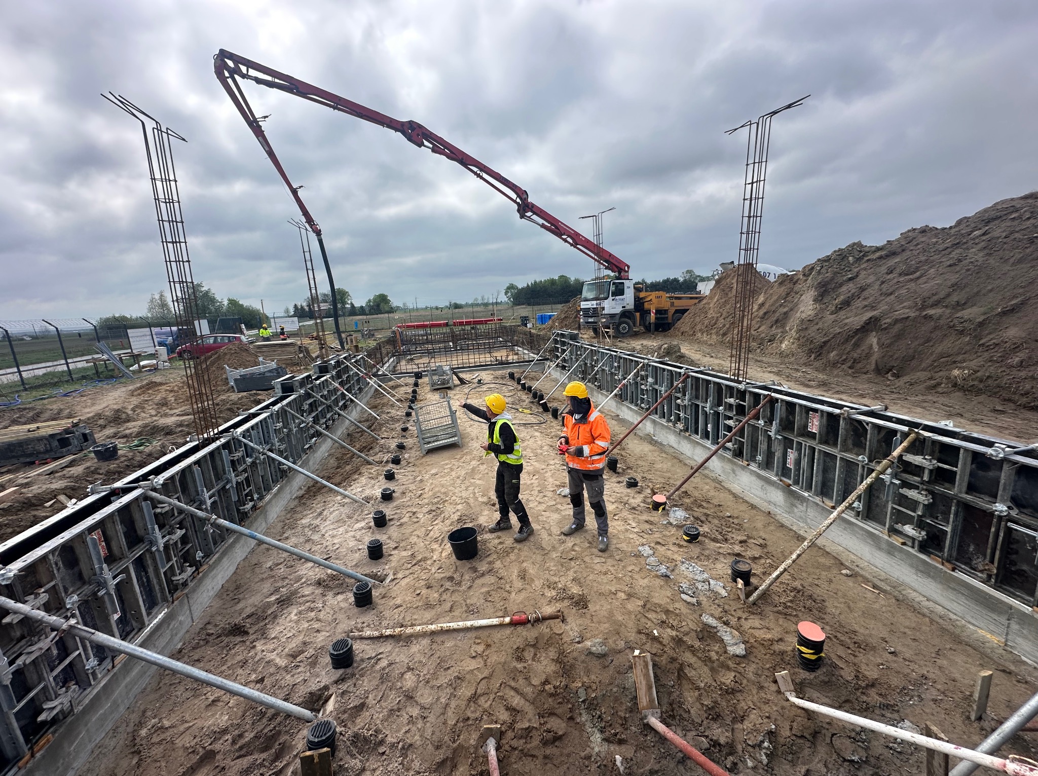Wykop z szalunkiem fundamentów, zbrojenie pionowe, dwaj pracownicy w kaskach i kamizelkach, pompa do betonu w tle, pochmurne niebo.
