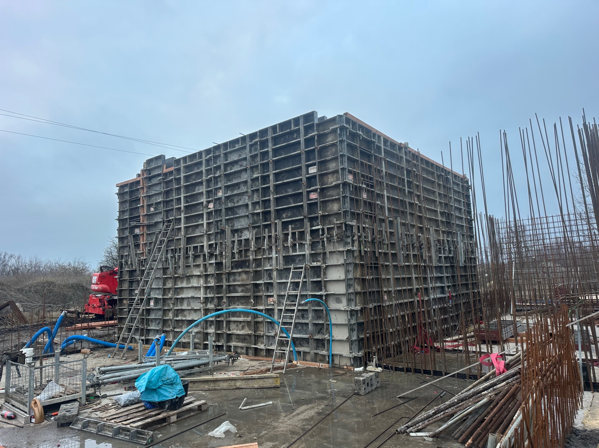 Szare szalunki fundamentowe z metalową konstrukcją i zbrojeniami na budowie w pochmurny dzień. Widoczne drabiny i węże doprowadzające beton.