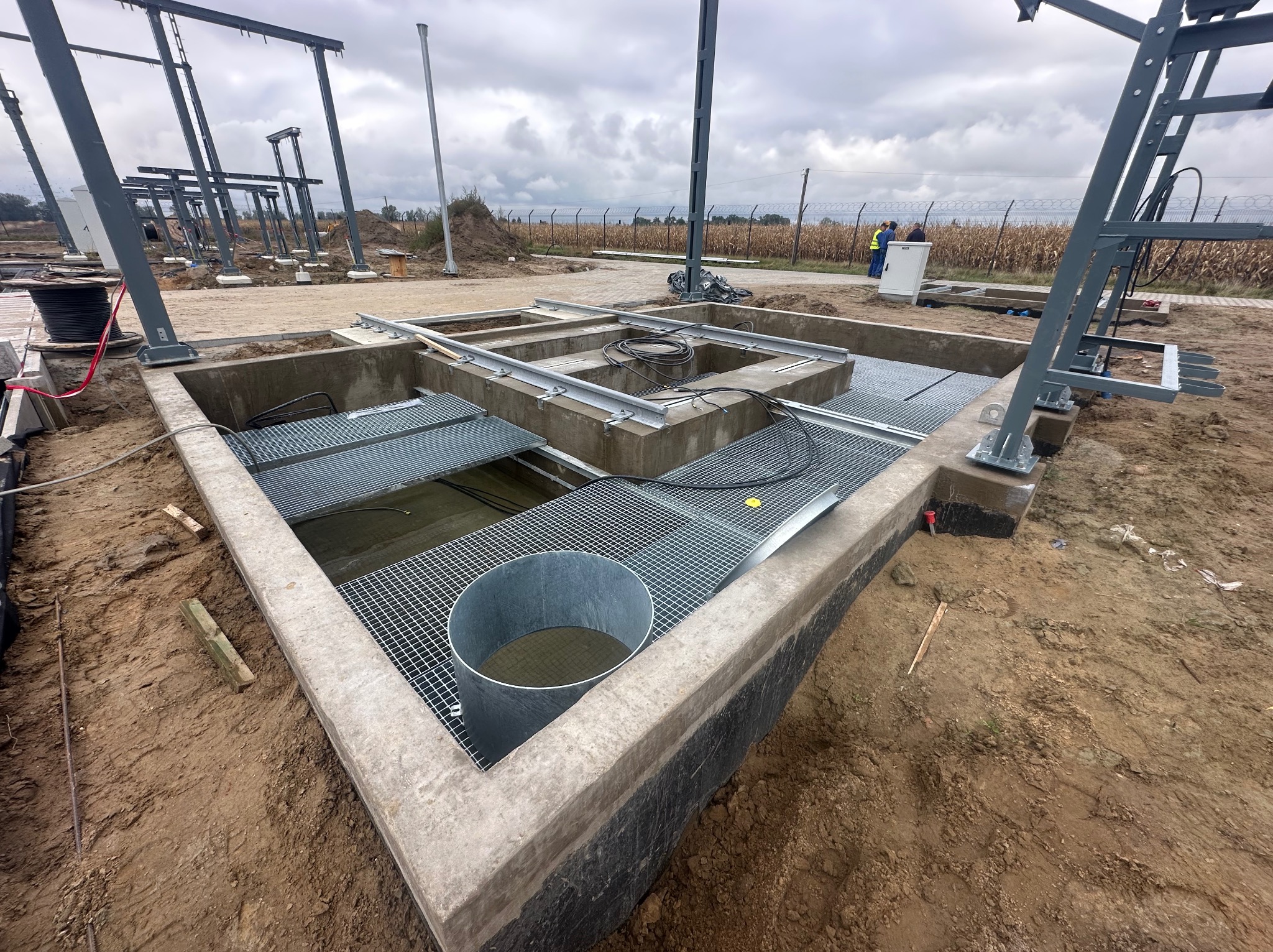 Wykop fundamentowy z betonowymi ścianami, metalowymi kratami i konstrukcją stalową. W tle widoczny płot z drutu kolczastego i pochmurne niebo.