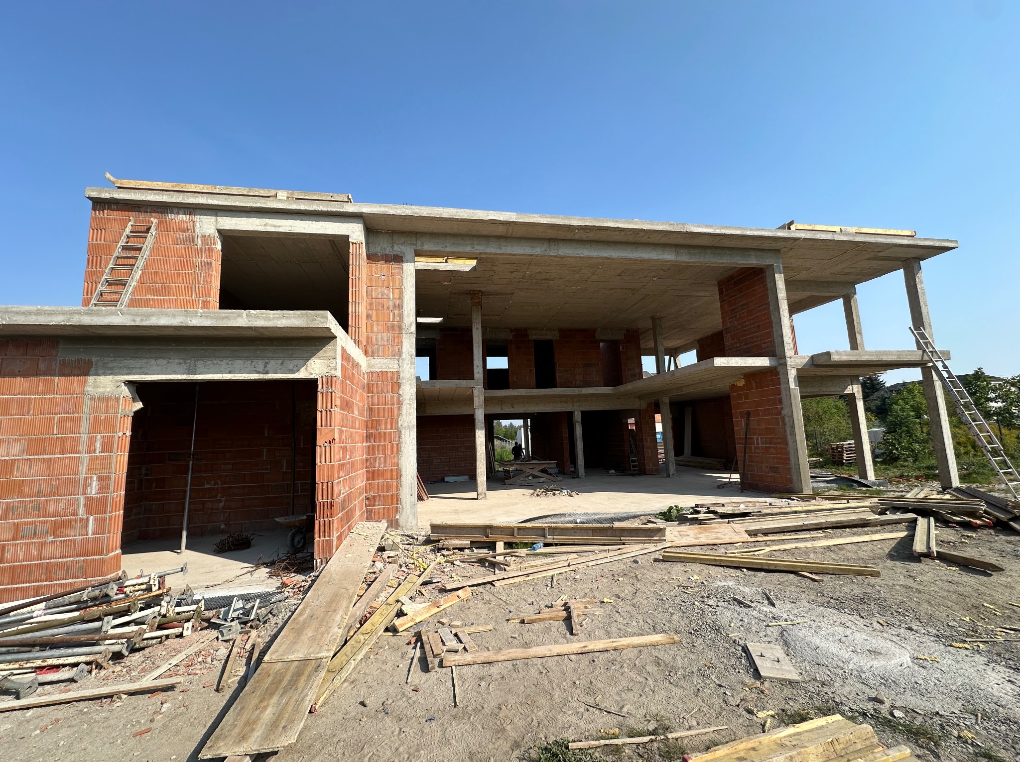 Surowy stan dwukondygnacyjnego domu z czerwonej cegły w trakcie budowy, z widocznymi elementami konstrukcyjnymi i rusztowaniami. Niebo błękitne w tle.