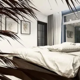 Minimalistyczna sypialnia z białymi tekstyliami na łóżku, czarną ramą drzwi i grafikami na ścianie, częściowo zasłonięta liśćmi palmy.