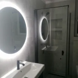 Nowoczesna łazienka z szarymi płytkami, okrągłym lustrem LED i kabiną prysznicową z czarnymi profilami, prezentująca minimalistyczny design i funkcjonalne rozwiązania.