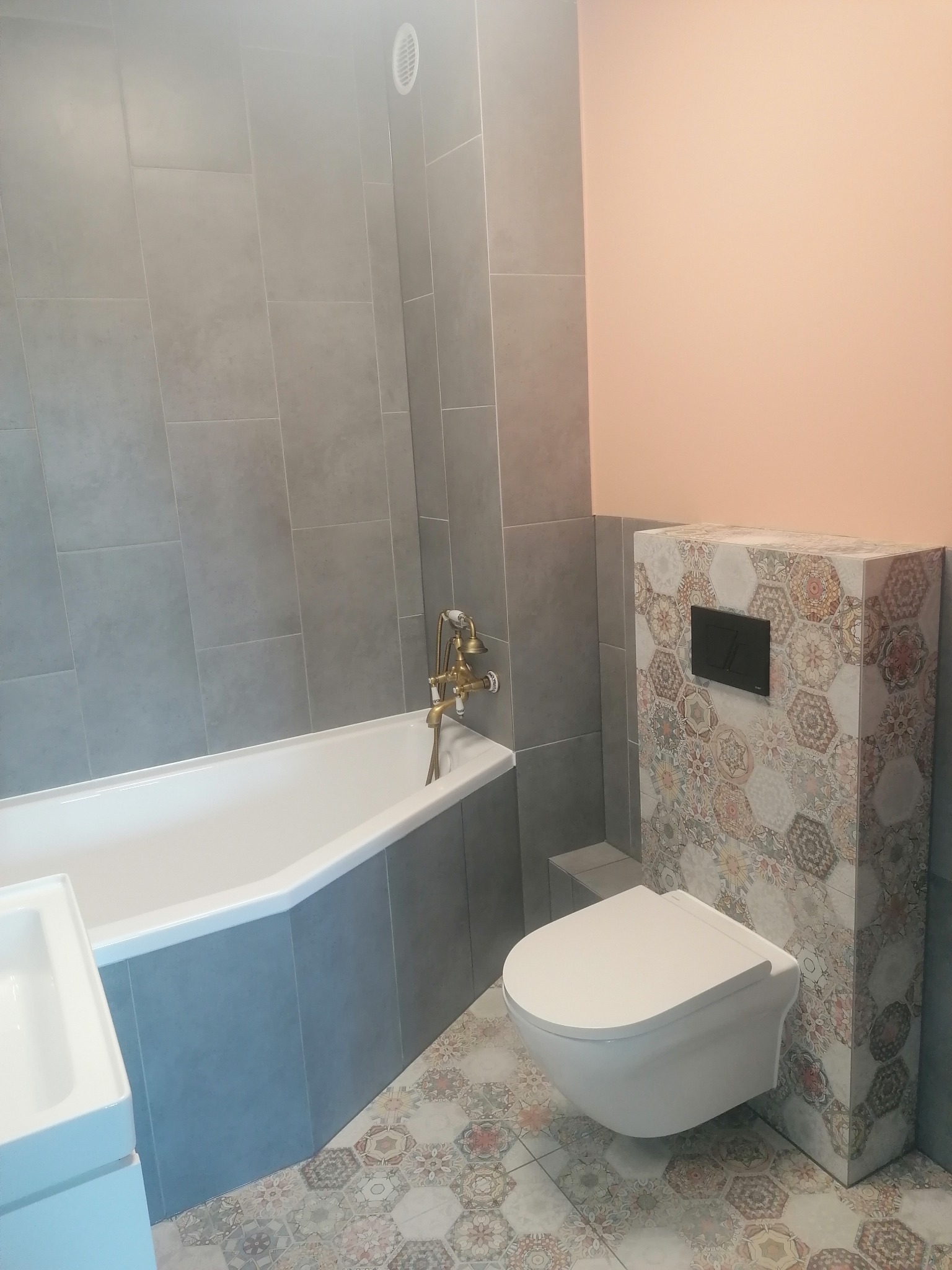 Nowoczesna łazienka z szarymi płytkami ściennymi, wanną z baterią w stylu retro, wiszącą toaletą z obudową w heksagonalne wzory i podłogą w pasujące wzory.