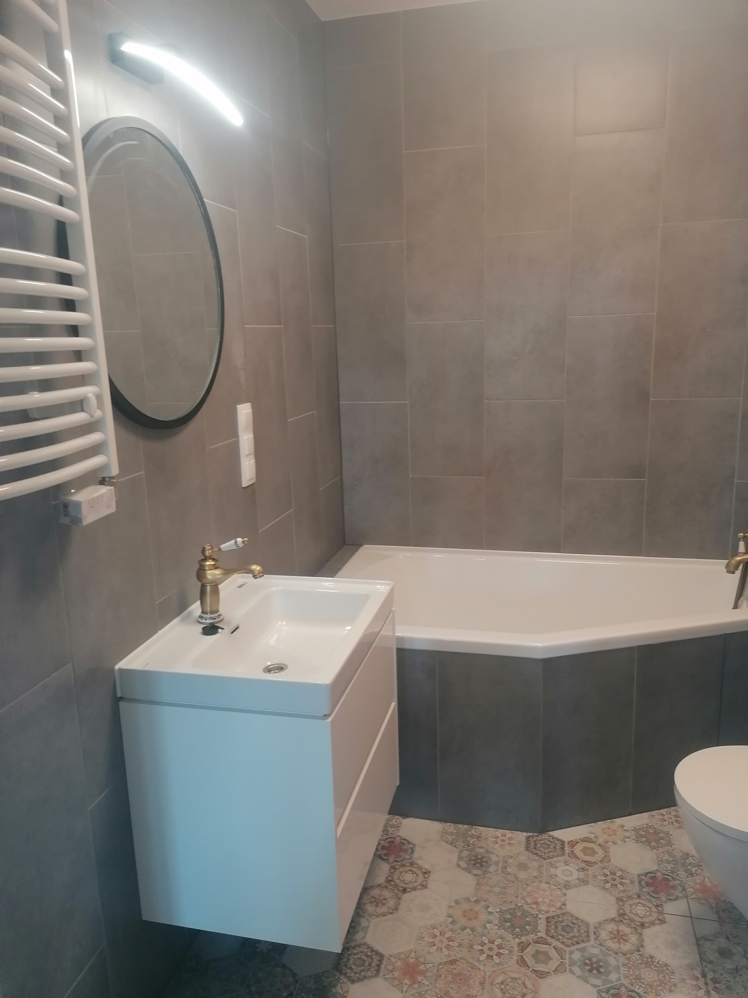 Wykończona łazienka z szarymi płytkami, białą umywalką z szafką, złotą armaturą, owalnym lustrem i dekoracyjnymi płytkami podłogowymi w stylu patchwork.
