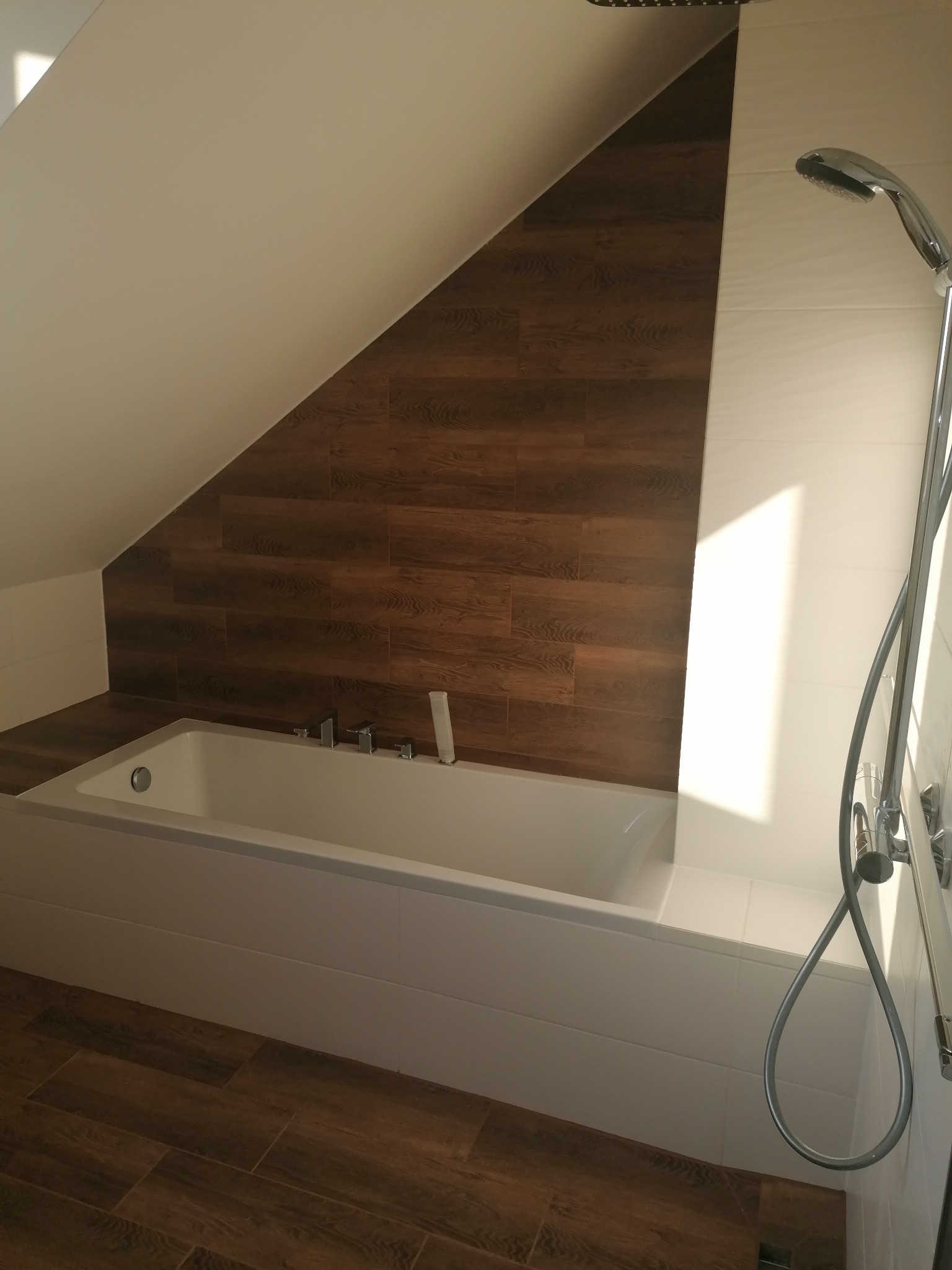 Nowoczesna łazienka na poddaszu z wanną obudowaną białymi płytkami, brązowymi płytkami imitującymi drewno na ścianie ze skosem oraz chromowaną słuchawką prysznicową.