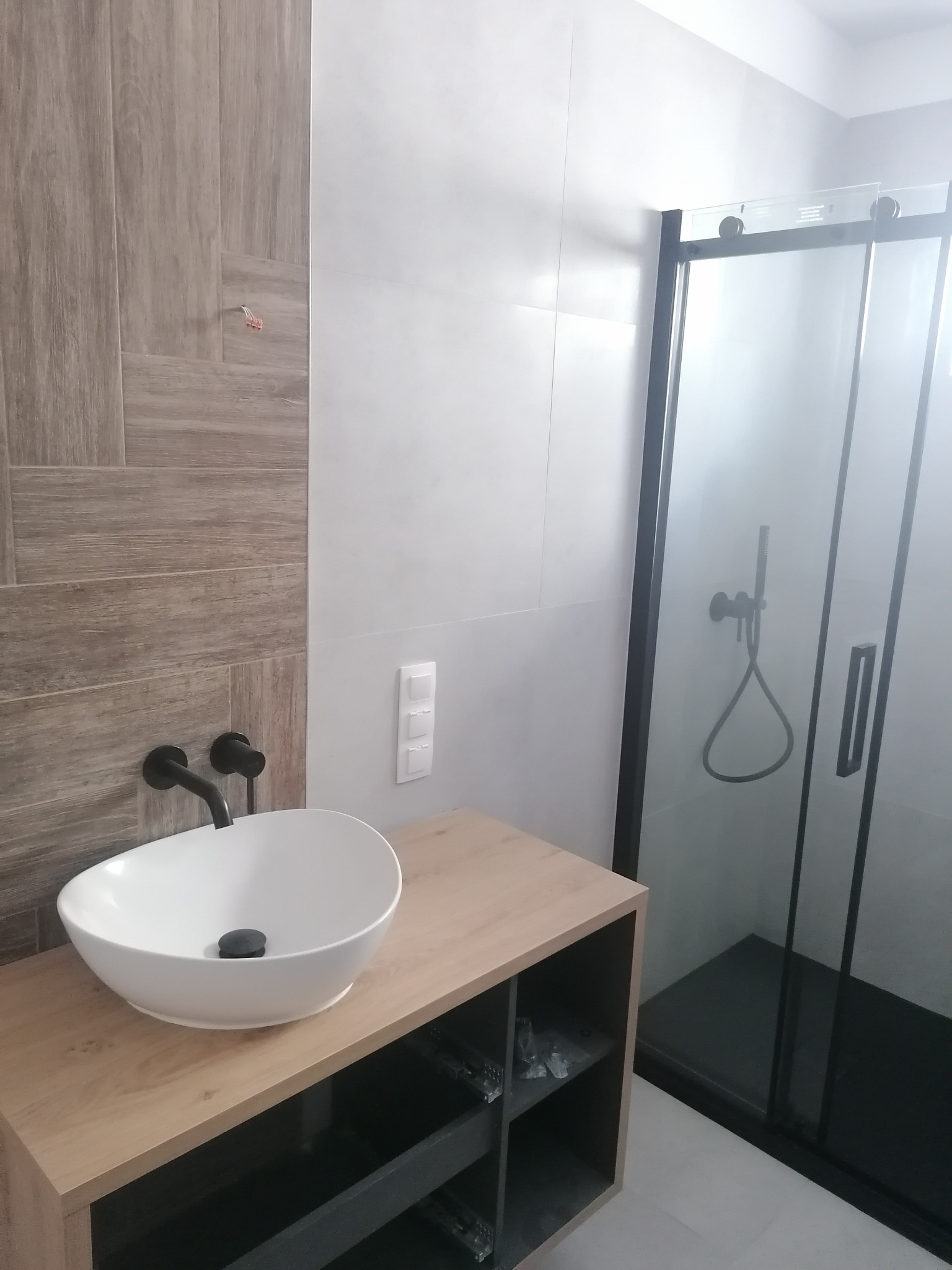 Nowoczesna łazienka z umywalką nablatową, czarną armaturą, kabiną prysznicową typu walk-in z czarnymi profilami i płytkami imitującymi drewno na ścianie.