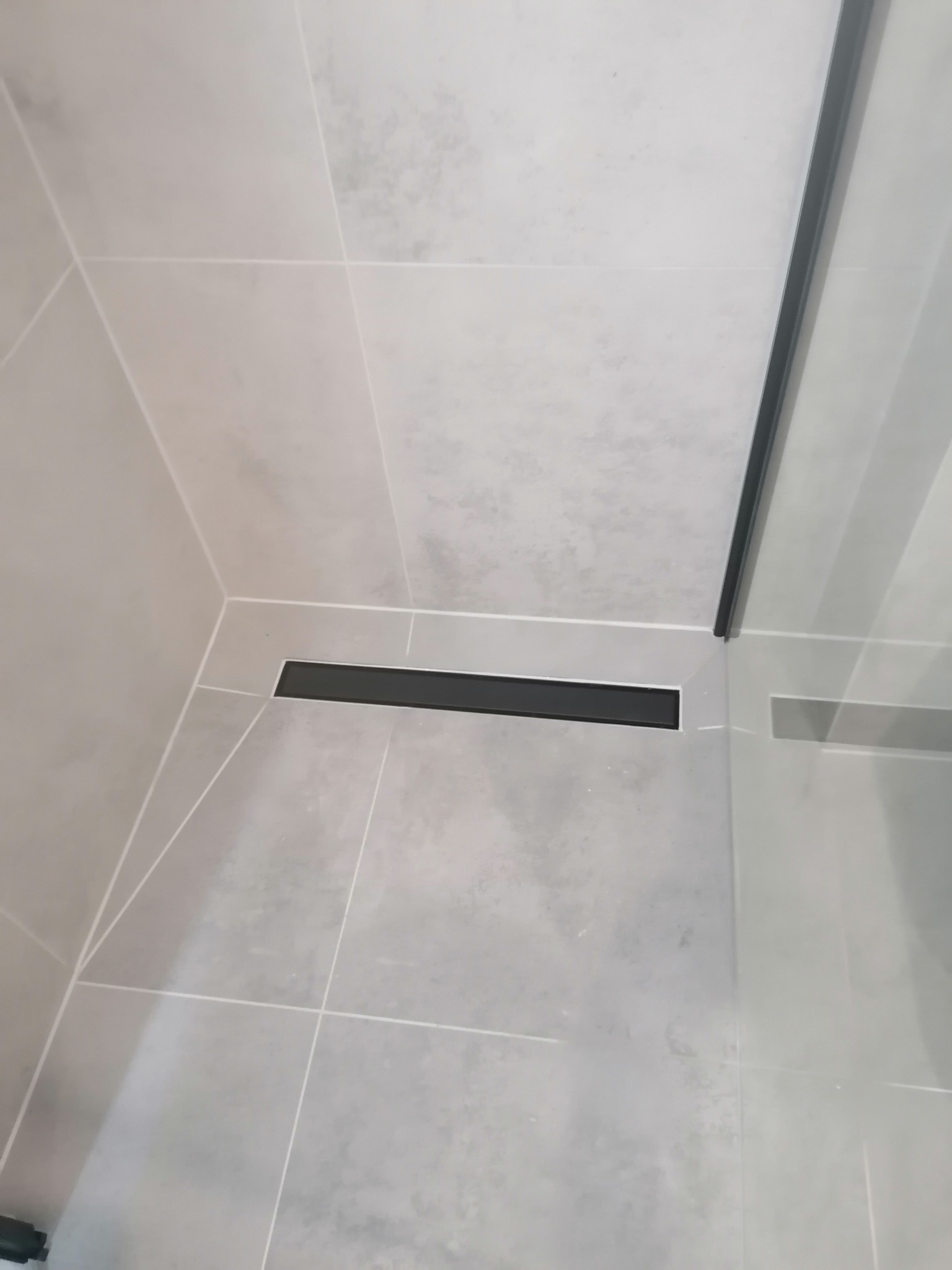 Szare płytki w kabinie prysznicowej z czarnym odpływem liniowym i szklaną ścianką.