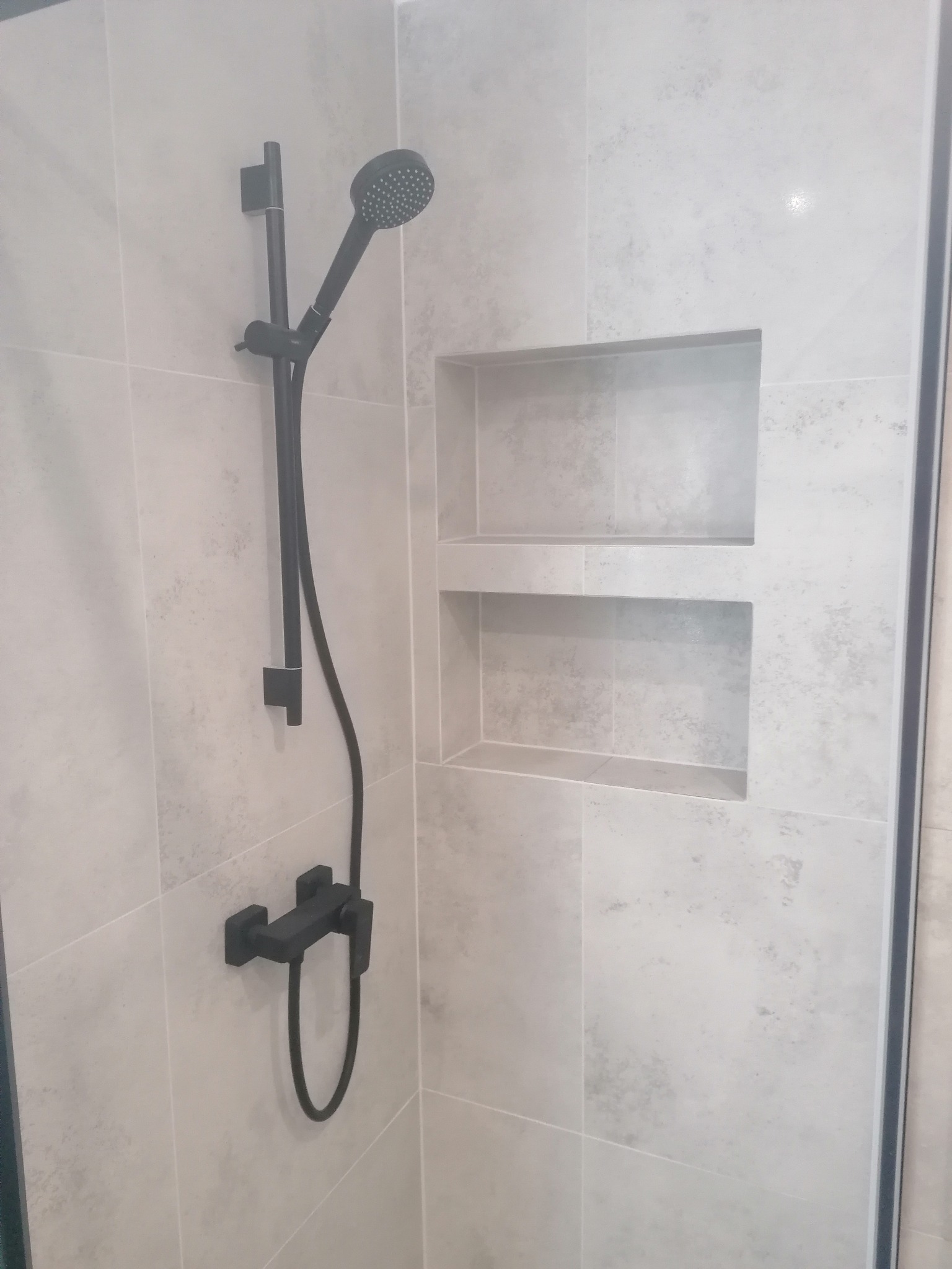 Nowoczesny prysznic z czarną armaturą na tle jasnoszarych, matowych płytek imitujących kamień, z wnękami na kosmetyki w ścianie.